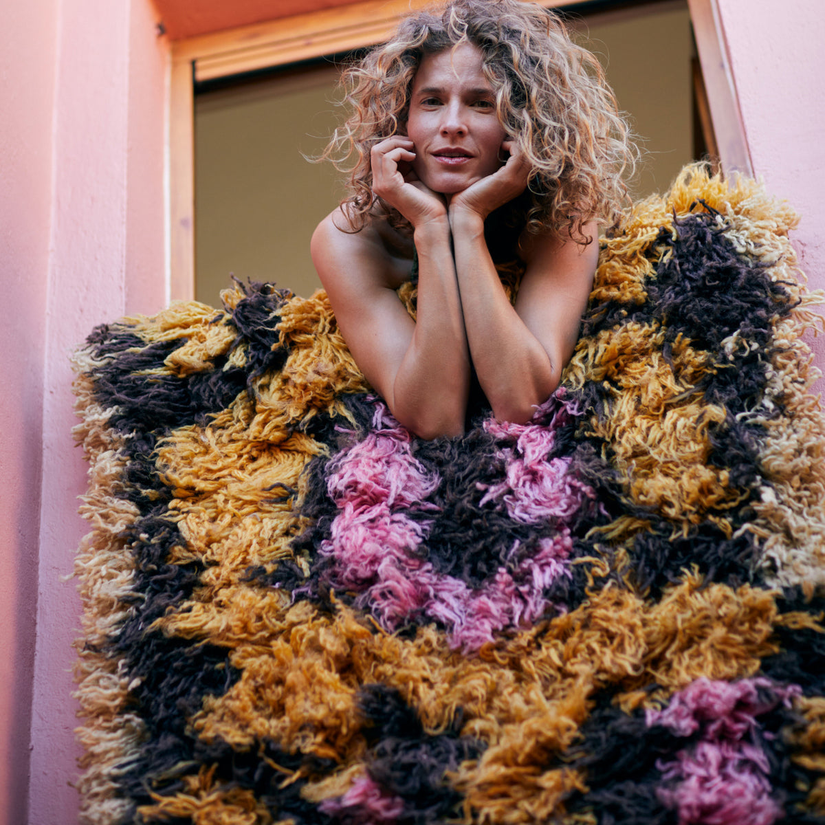 Een vrouw met krullend haar glimlacht terwijl ze op een vensterbank leunt, haar armen rustend op de HK Living Fluffy Tapijt South Beach 360/80, met gele, roze en zwarte patronen. Roze muren en een open raam maken het vrolijke tafereel compleet.