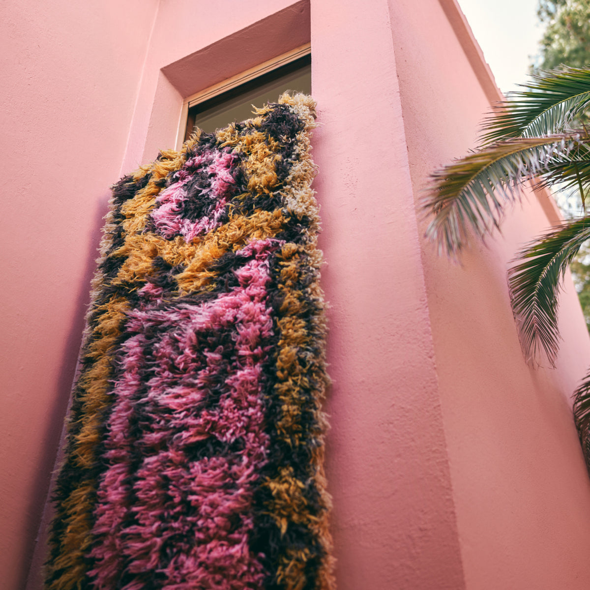 De HK Living Fluffy Tapijt South Beach 360/80 in roze, bruin en geel hangt uit een raam van een roze gebouw, met rechts een palmboom en een lichte lucht, wat de speelse stijl van HK Living weerspiegelt.