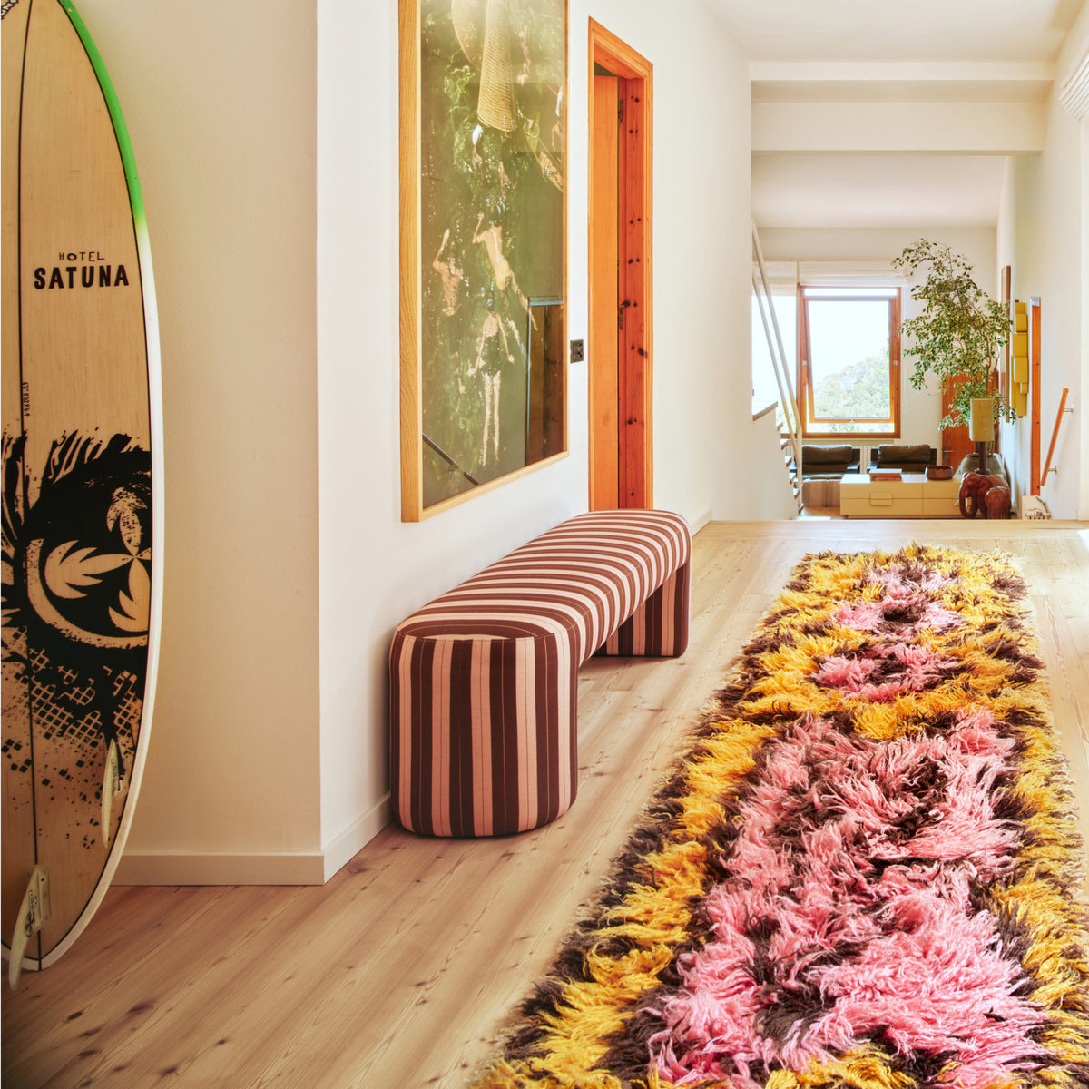 Een lichte hal is voorzien van een gestreepte bank, een HK Living surfplank, het HK Living Fluffy Tapijt South Beach 360/80 vloerkleed, grote kunstwerken en een doorkijk naar het zonnige woongedeelte met groen buiten.