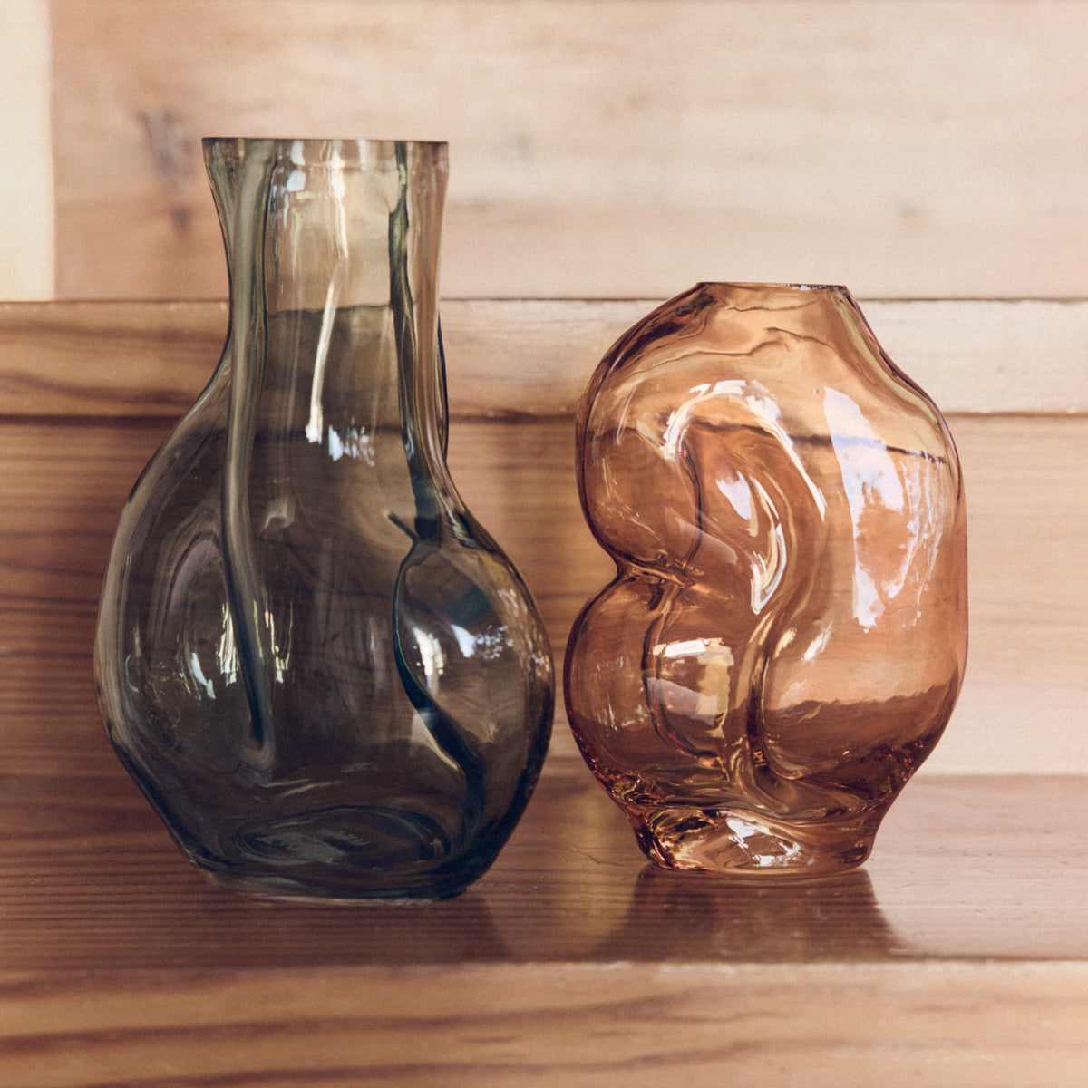 De HK Living Gleam Vaas Olijfgroen H22, een abstracte gebogen glazen vaas in olijfgroen, staat op een houten ondergrond tegen een natuurlijke houten achtergrond.