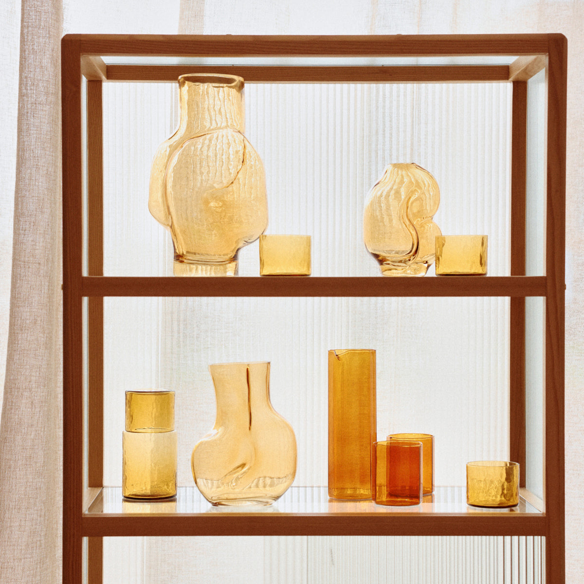 Op een houten plank staat de HK Living Gleam Vaas Amber H18 van HK Living, samen met andere glazen vazen in verschillende vormen en maten, tegen een lichte, doorschijnende achtergrond.