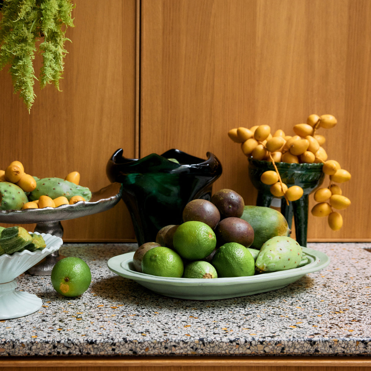 Een aanrecht toont groen en geel fruit - limoenen, cactusvijgen, dadels en passievruchten - gerangschikt in de HK Living New Classics Schaal Muntgroen L. Daarboven hangt groen met een donkere vaas tegen houten kasten.