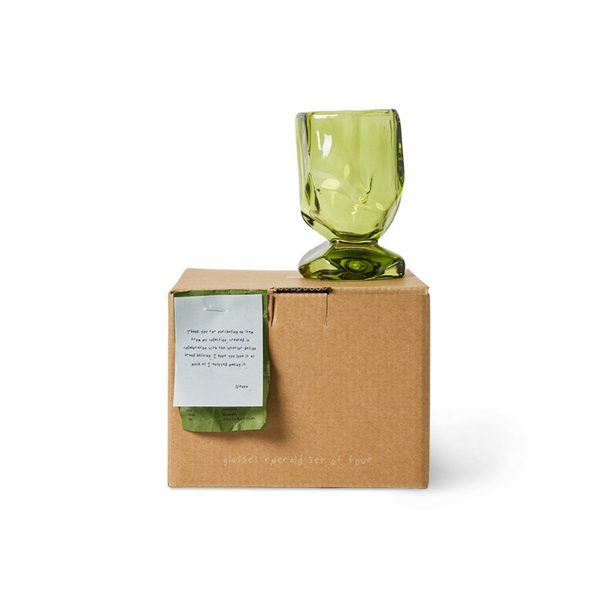 Een set van vier HK Living Nienke Sikkema-wijnglazen in de kleur Emerald Groen, met een uniek groen ontwerp, staat op een bruine doos waarop het product en het merk staan vermeld, met daarbovenop een handgeschreven briefje dat een van de vier glazen gedeeltelijk bedekt, tegen een witte achtergrond.