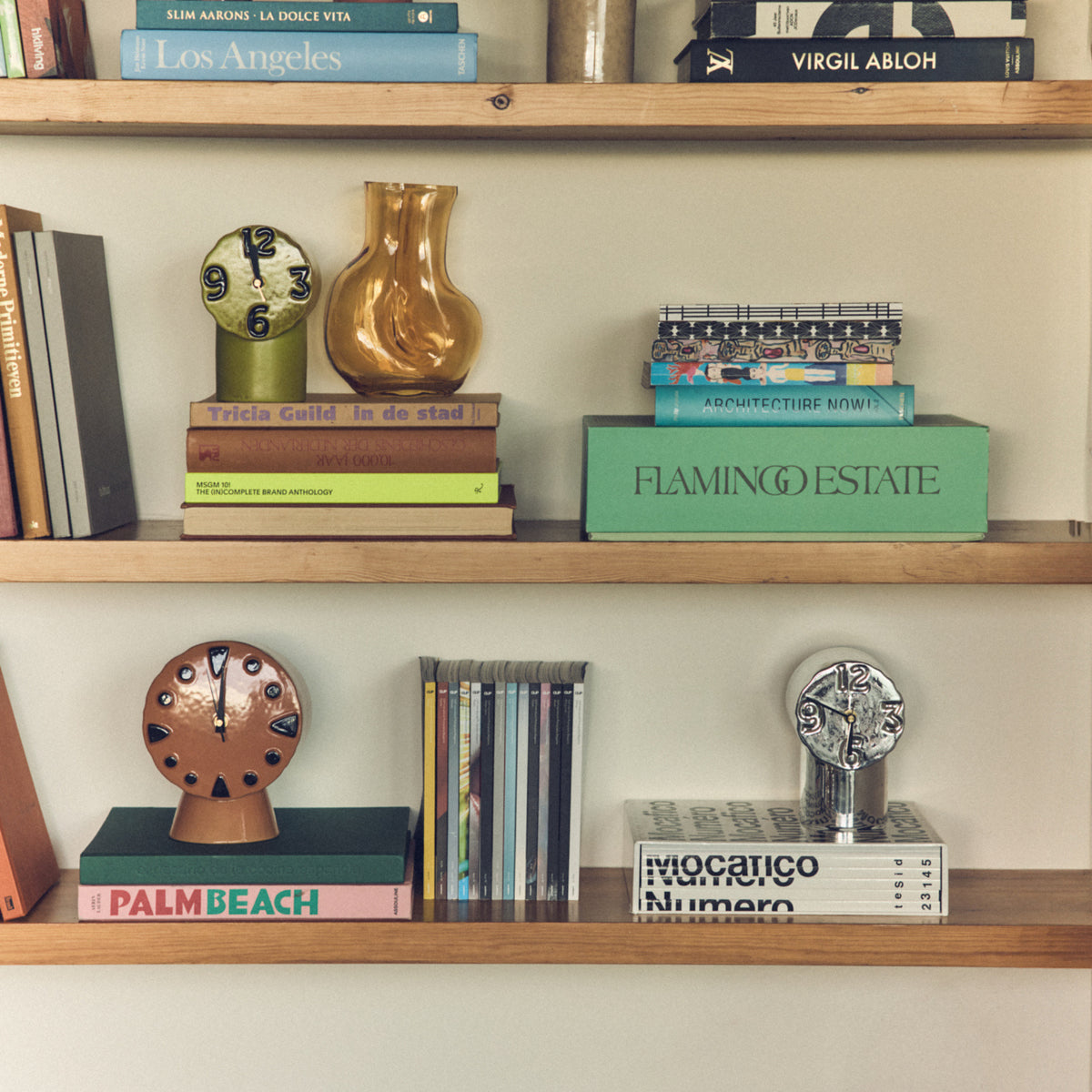 Op een netjes geordende boekenplank staan kleurrijke boeken, kunsttijdschriften, kleine HK Living Retro Klok Chroom klokken, een gele glazen vaas, een organische vaas en een stapel tijdschriften op houten planken.