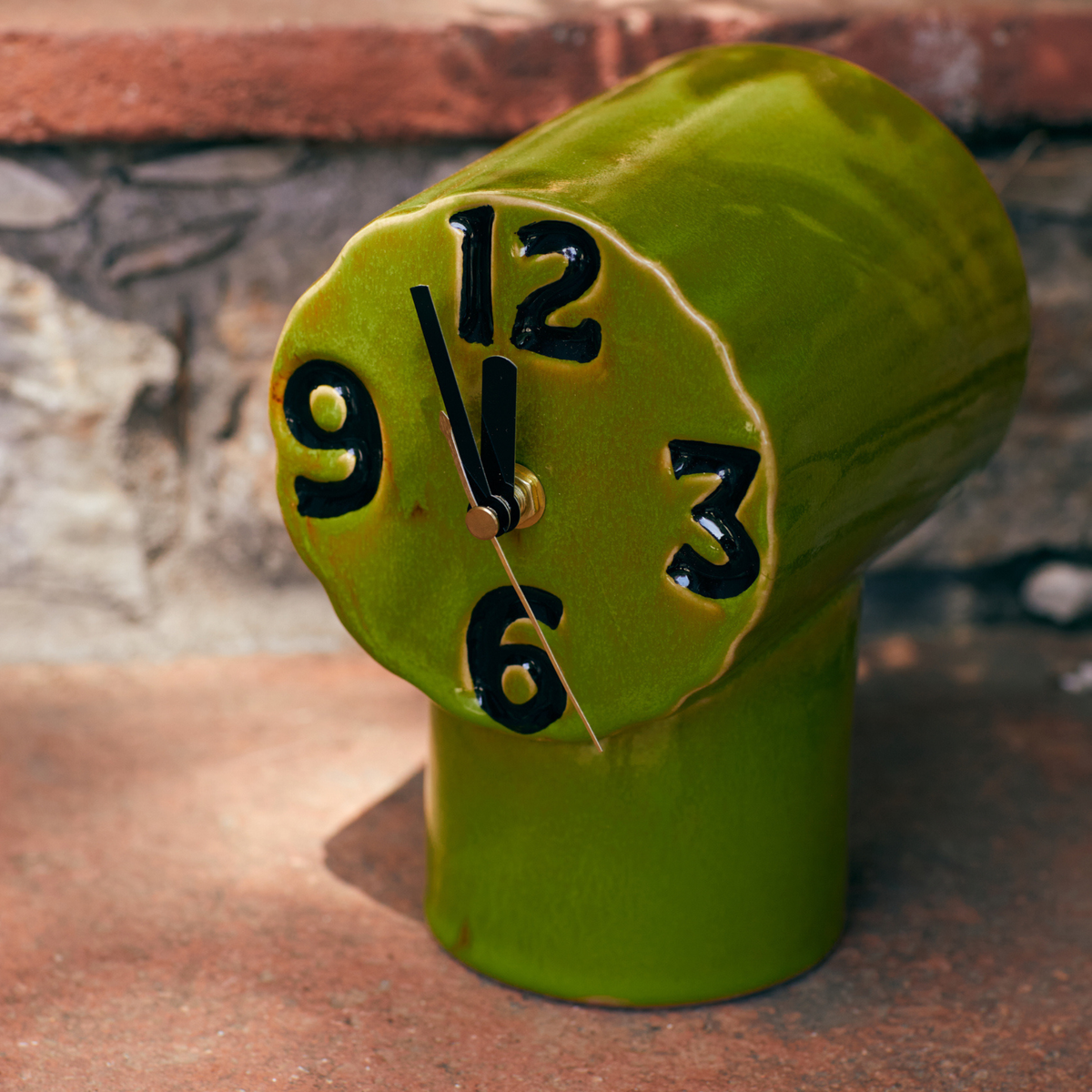 De HK Living Retro Klok Olijfgroen, met zijn unieke, vloeiende keramische vorm, opvallende zwarte cijfers en gouden wijzers die op 11:00 uur staan, staat op een stenen ondergrond tegen een stenen muur.