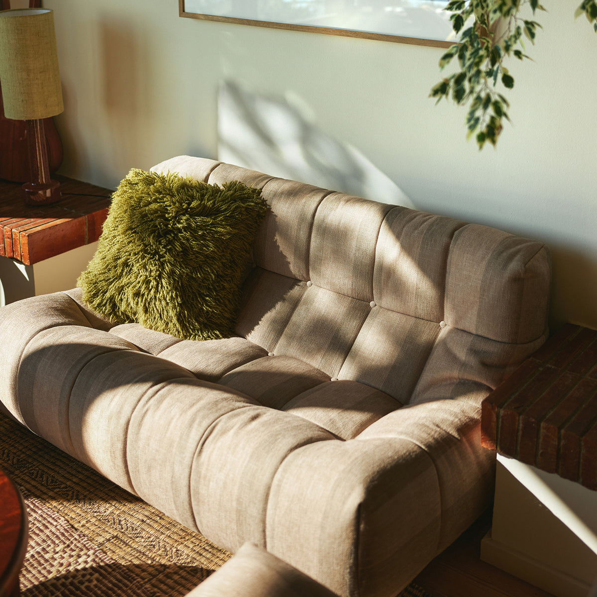 Een beige getufte loveseat met een HK Living Rugged Kussen Olijfgroen 50/50 kussen staat in een zonovergoten hoek, naast houten bijzettafeltjes en een hangende bladplant.