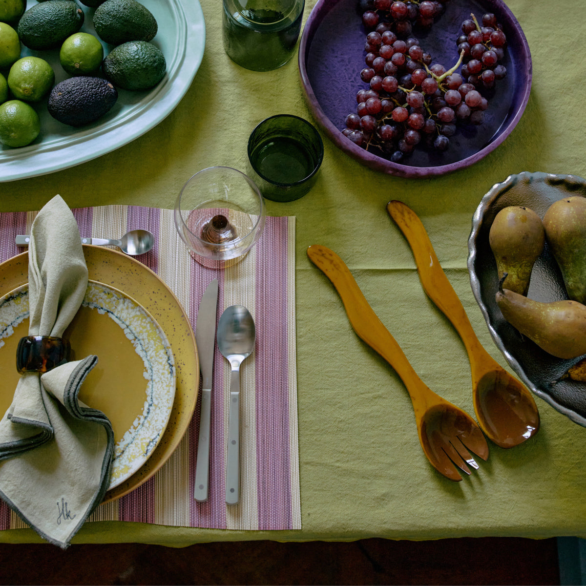 Een gedekte tafel met limoenen, avocado's, rode druiven, peren, servet en bestek, twee groene glazen en HK Living Saladebestek Oker 2 st. op een groen tafelkleed-perfect als attent cadeau van HK Living.