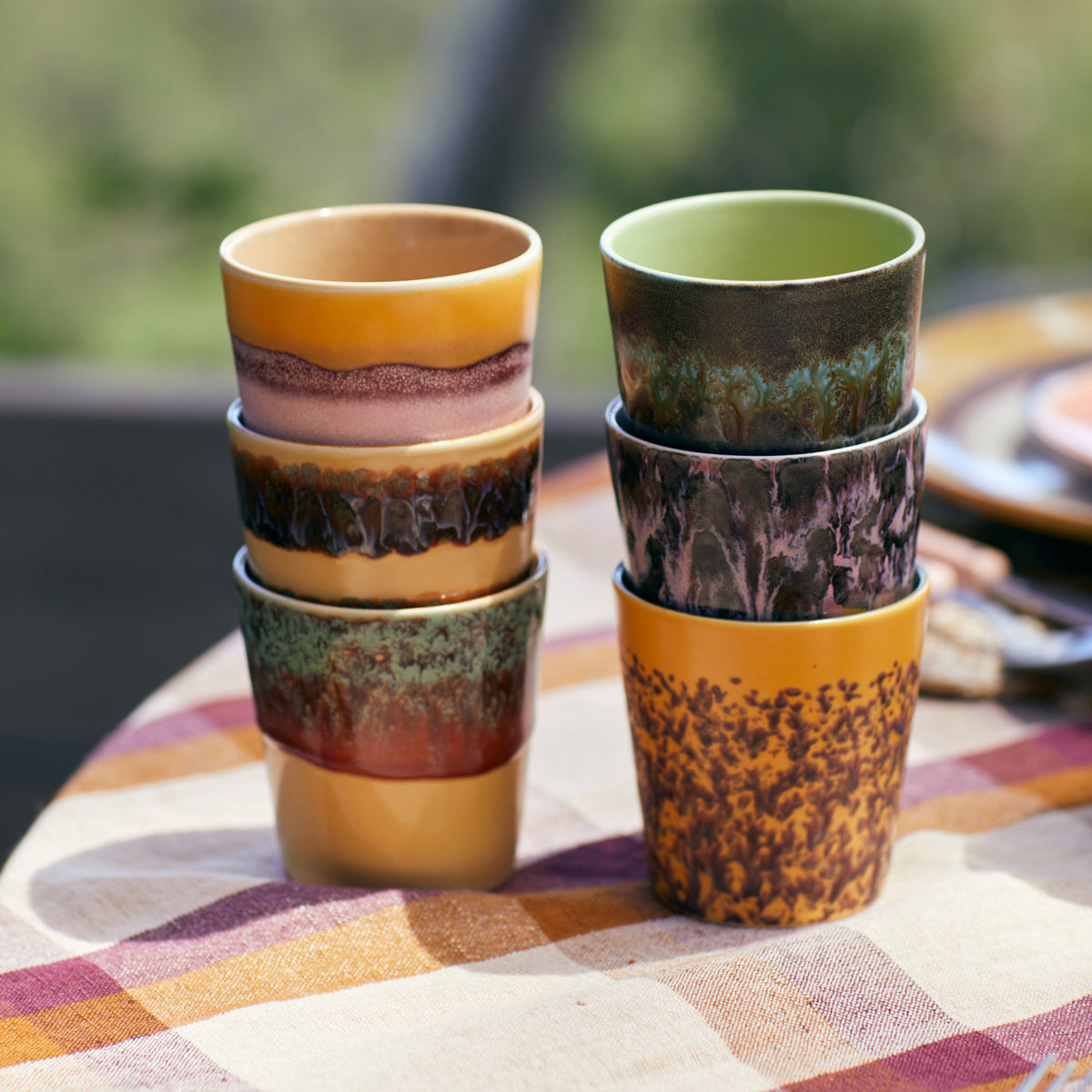 Zes HK Living 70s Ceramics Koffiekop Cove-kopjes, elk met een uniek glazuurpatroon, zijn paarsgewijs gestapeld op een geruit tafelkleed buiten en brengen retrostijl in je omgeving.