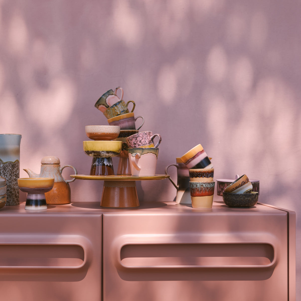 Een stapel HK Living 70s Ceramics Koffiekop Cliffs kopjes staat op een roze kast tegen een roze muur, met zacht gedimd zonlicht dat retro stijl schaduwen toevoegt aan het kleurrijke tafereel.