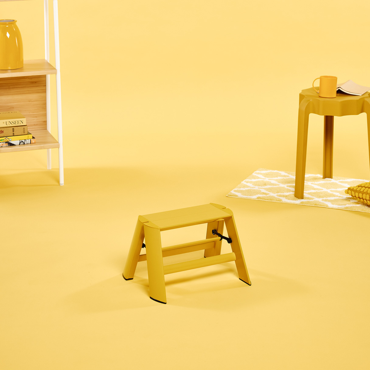 Een Hasegawa Lucano Ladder 1 Step Geel van Hasegawa, geïnspireerd op een minimalistisch ontworpen opstapje, staat op een gele vloer in een kamer met een gele tafel, een mok, een pot en een plank met boeken; ook de achtergrond is geel.