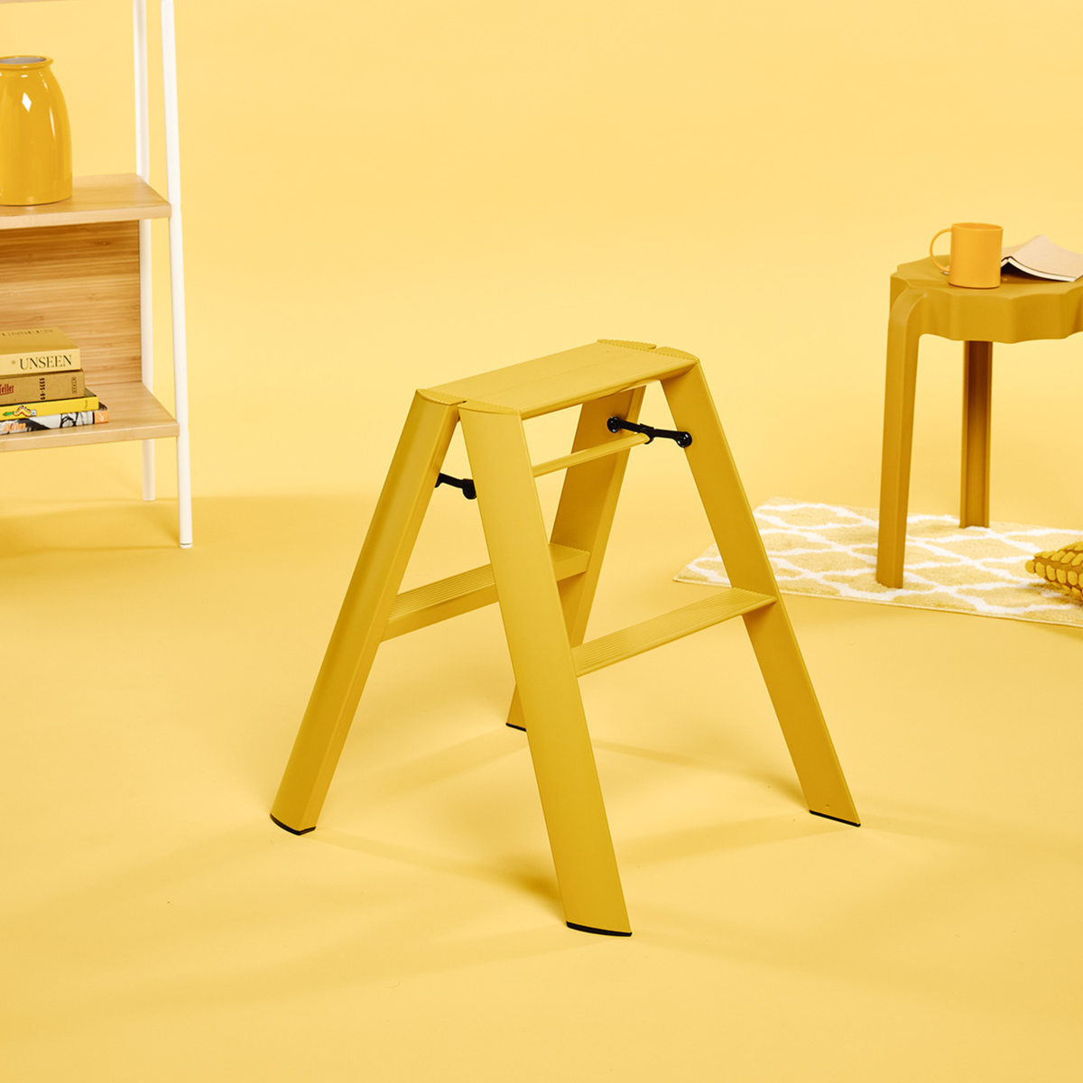 Een Hasegawa Lucano Ladder 2 Step Geel van Hasegawa staat in een kamer waar de muren, de vloer en het meubilair – waaronder een tafeltje met een kopje en boeken op een plank – allemaal geel zijn, waardoor een monochroom geel thema ontstaat dat is geïnspireerd op Japans design.