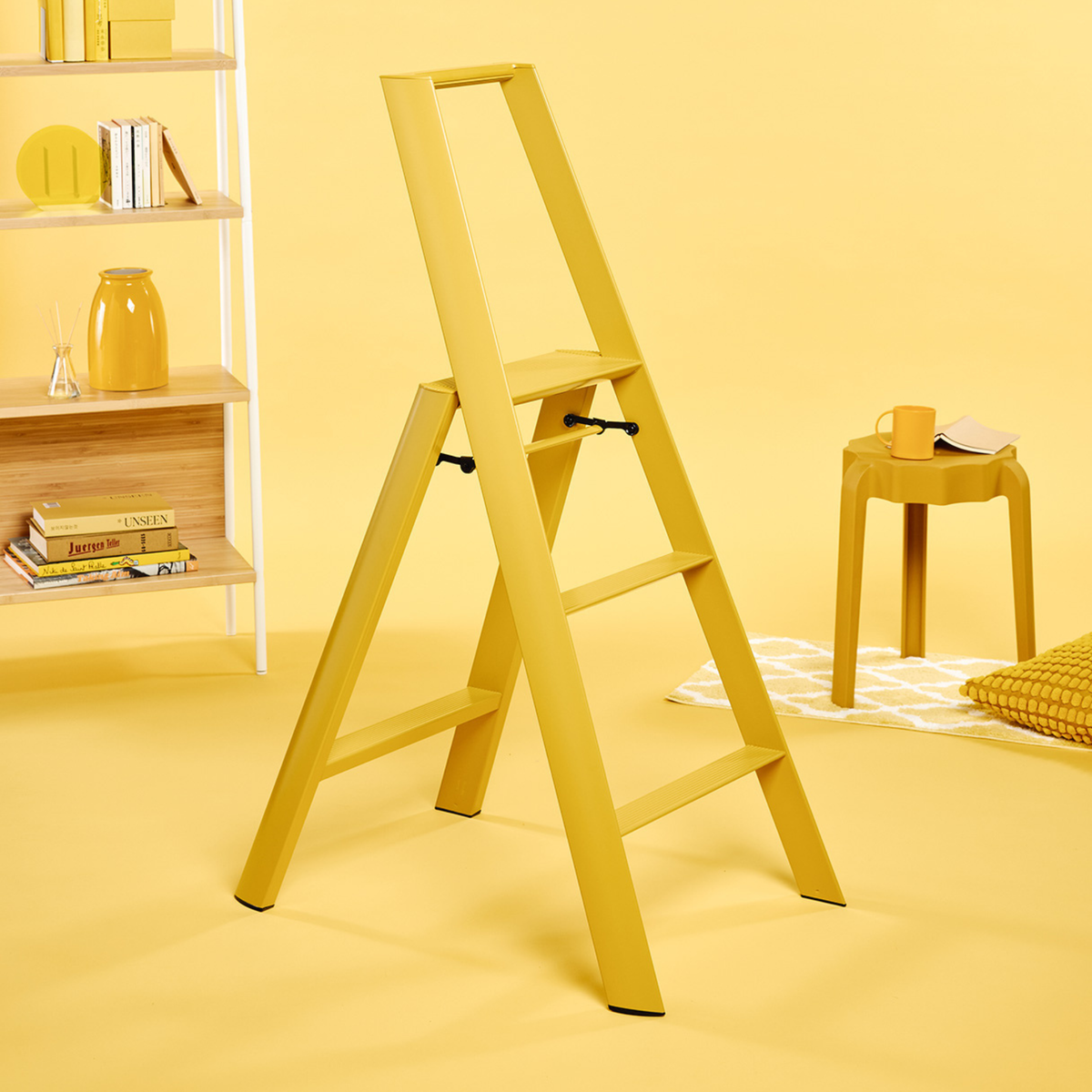 Een Hasegawa Lucano Ladder 3 Step Geel van Hasegawa staat op een gele vloer, met op de achtergrond een bijpassend geel krukje, vloerkleed en boekenkast, wat samen een monochroom tafereel vormt dat is geïnspireerd op de Japanse ontwerpesthetiek.
