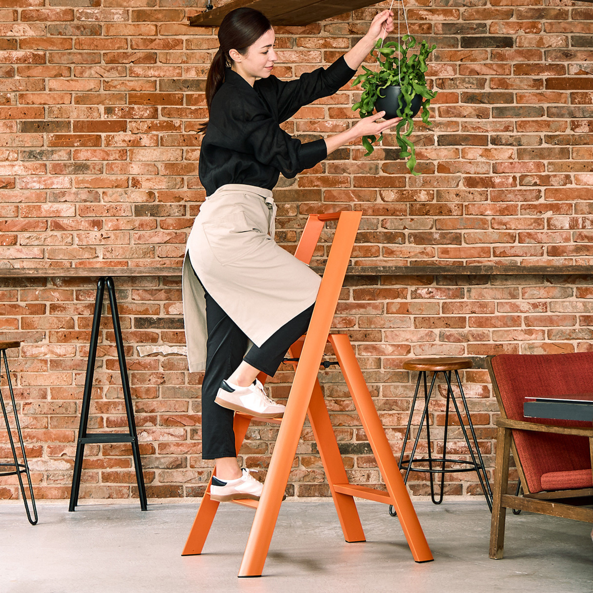 Een vrouw in een zwart shirt en een beige schort staat op een Hasegawa Lucano Ladder 3 Step Oranje van Hasegawa en zet een hangplant recht tegen een bakstenen muur in een moderne, gezellige kamer.