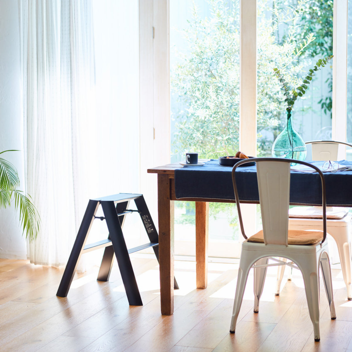 Lichte eethoek met houten tafel, witte metalen stoelen, een Hasegawa Lucano Ladder 2 Step Zwart van Hasegawa en een grote glazen deur die zonlicht binnenlaat en groen buiten onthult; een heldere vaas op tafel voegt een Japanse designflair toe.
