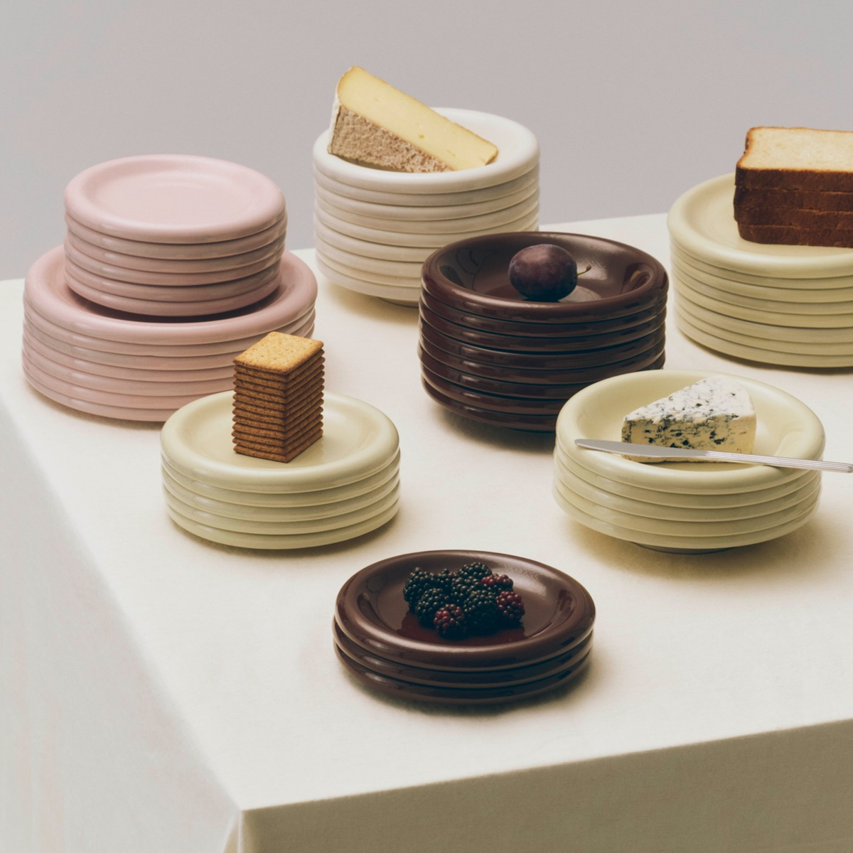 Stapels pastelkleurige en donkere borden, waaronder de Hay Barro Borden Bordeaux D18 2 st. van Hay, staan op een crèmekleurig tafelkleed, elk belegd met kaas, brood, bessen, crackers en een chocoladetruffel tegen een neutrale achtergrond.