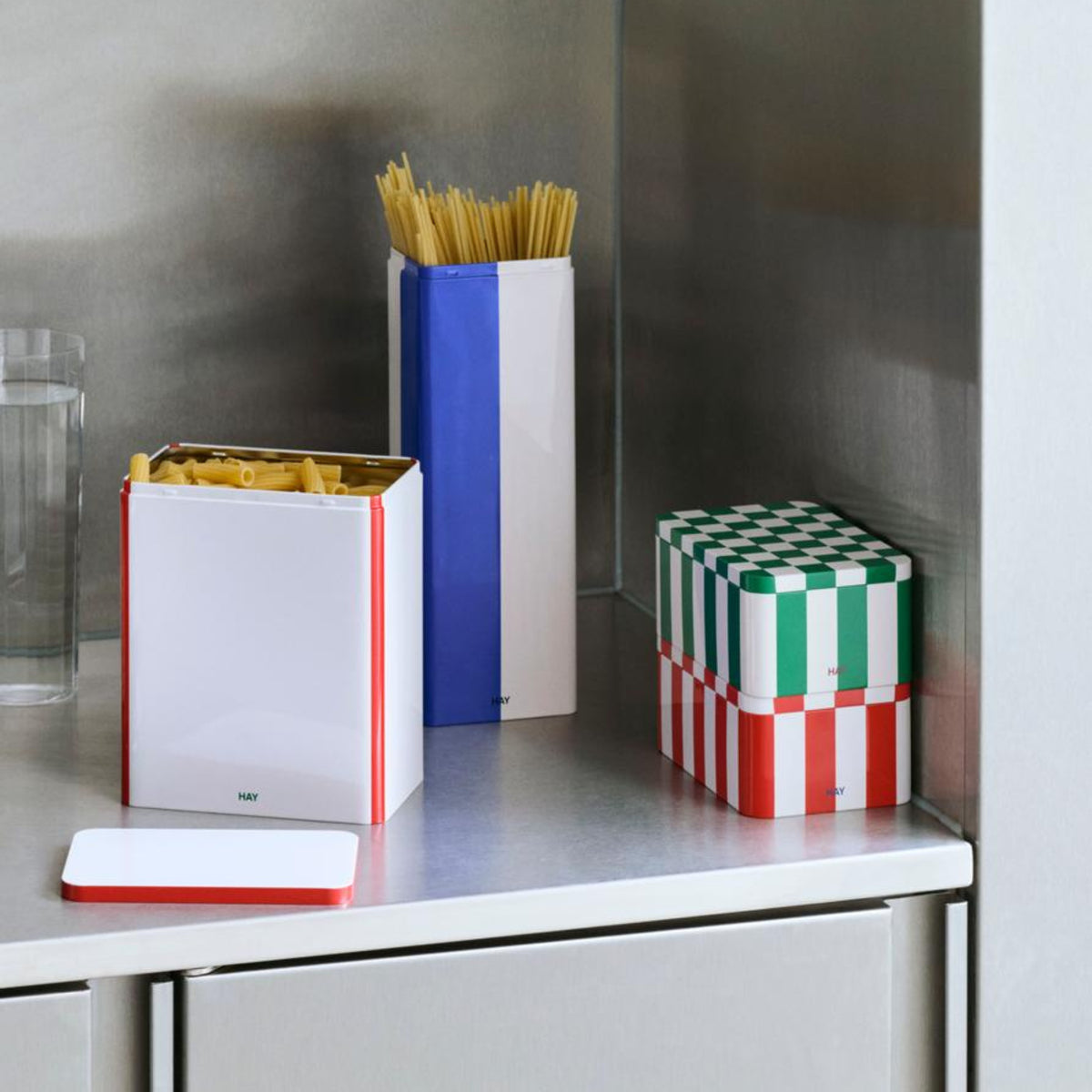 Een aanrecht met een glas water, ongekookte spaghetti, een open pastabak en twee gestapelde Hay Blikken Doos Blauw Off-White Slim voorraadblikken voegt een vleugje Scandinavisch design toe aan het roestvrij stalen oppervlak.