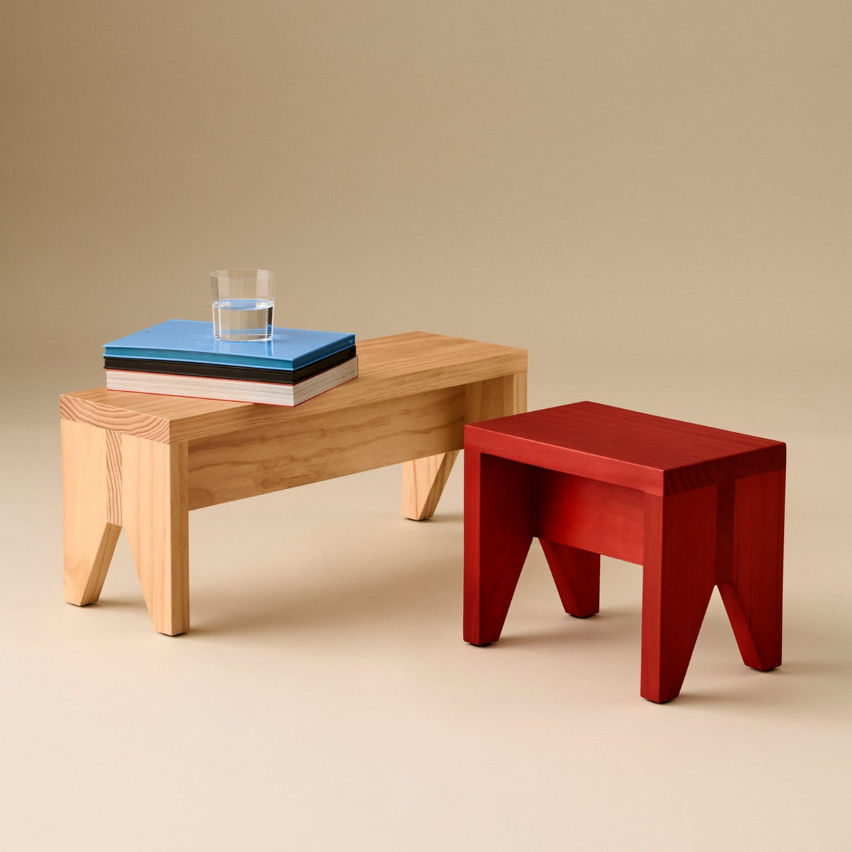 Twee kleine Hay Manolito Stool Bank Naturel Lang krukjes staan op een beige achtergrond; op het lichthouten krukje staan boeken en een glas water, terwijl de rode versie iets naar voren is geplaatst.