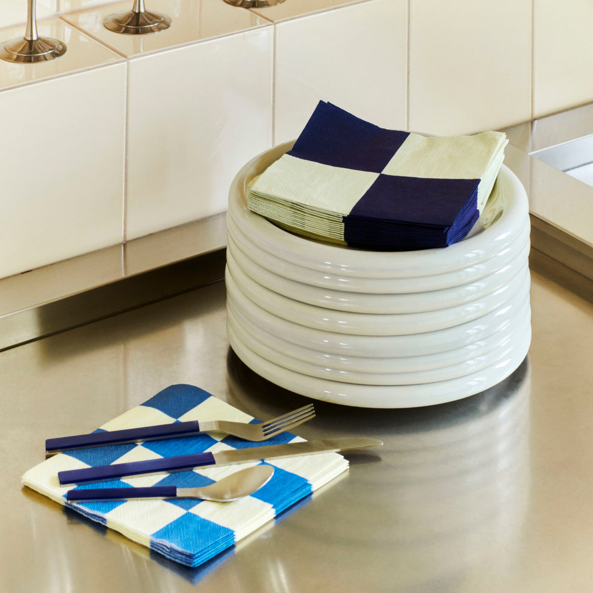 Een stapel witte borden met Hay Pattern Papieren Servetten Off-white Blauw Ruit 33/33 van Hay staat op een roestvrijstalen aanrechtblad, naast bijpassende servetten met blauw bestek. Wit betegelde achterwand op de achtergrond.