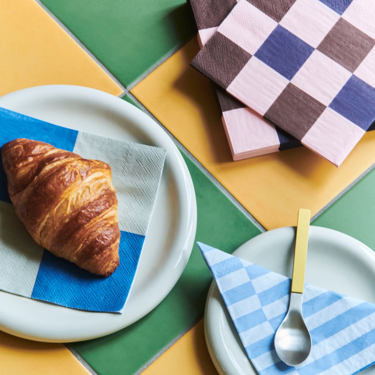 Een croissant op een blauw en wit servet op een bord, een lepel met een blauw geruit servet en twee gestapelde Hay Pattern Papieren Servetten Roze Bordeaux Zwart 33/33 van Hay op gele en groene tegels.