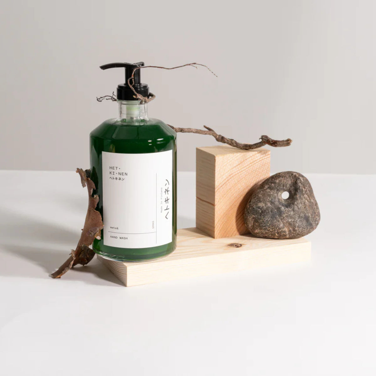 De Hetkinen Handzeep Metsa 500 ml van Hetkinen, met een zwarte pomp en groene fles, rust op een licht houten blok naast een gladde steen en takjes gemaakt met biologische plantaardige oliën voor een eco-vriendelijke vibe tegen een neutrale achtergrond.