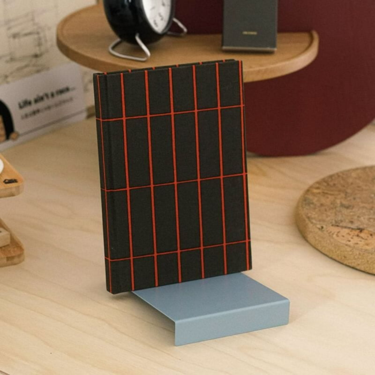 Een zwart notitieboek met een rood rasterpatroon staat rechtop op de Hightide Boekenstandaard & Display Lichtblauw van Hightide, geplaatst op een houten bureau dat is bezaaid met kantoorartikelen zoals een klok, papieren en een kurkmat.