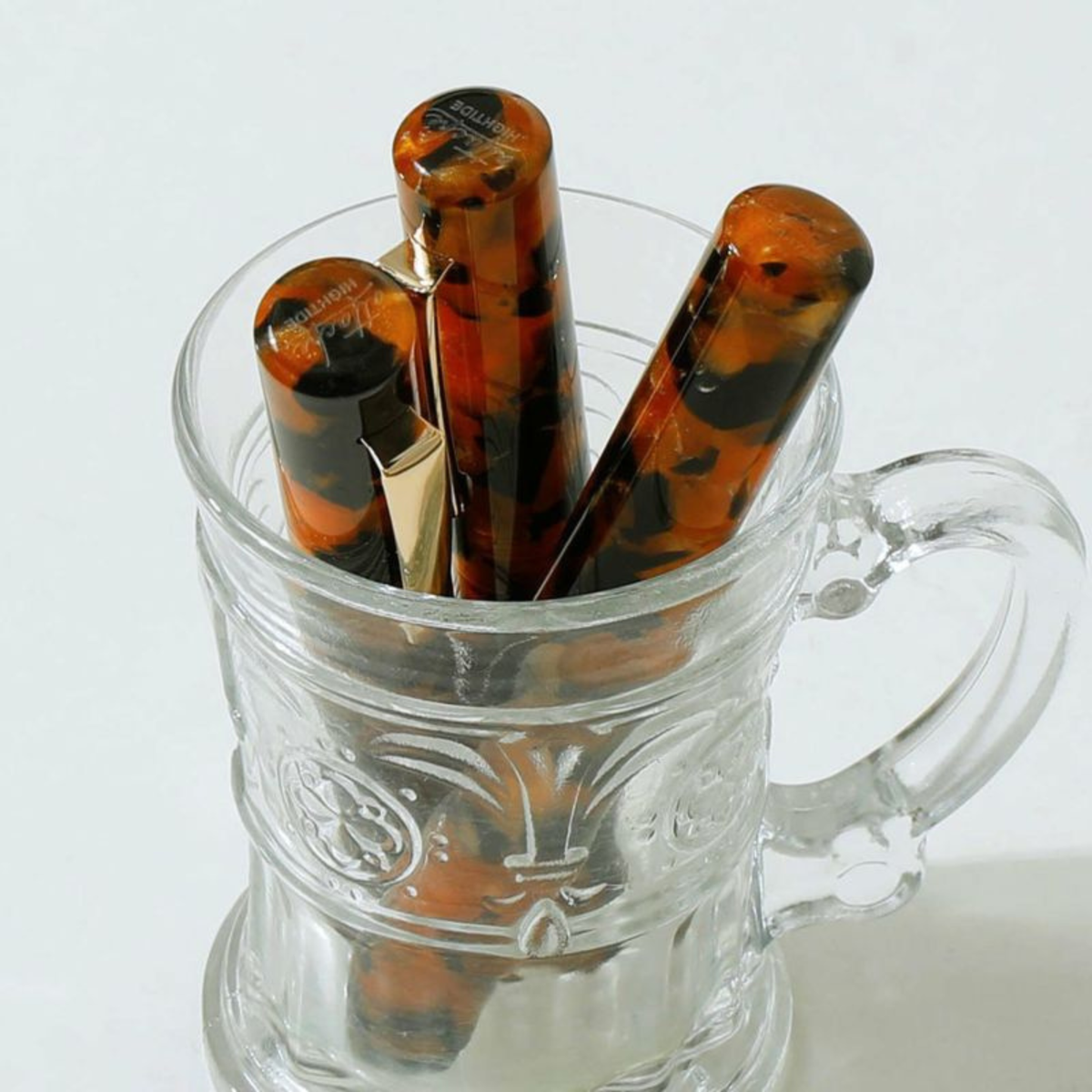 Drie Hightide Marmeren Fountain Vulpen Bruin-pennen, met een bruin gemarmerd patroon en gouden clips, staan opgesteld in een doorzichtige glazen beker met een handvat, tegen een witte achtergrond.