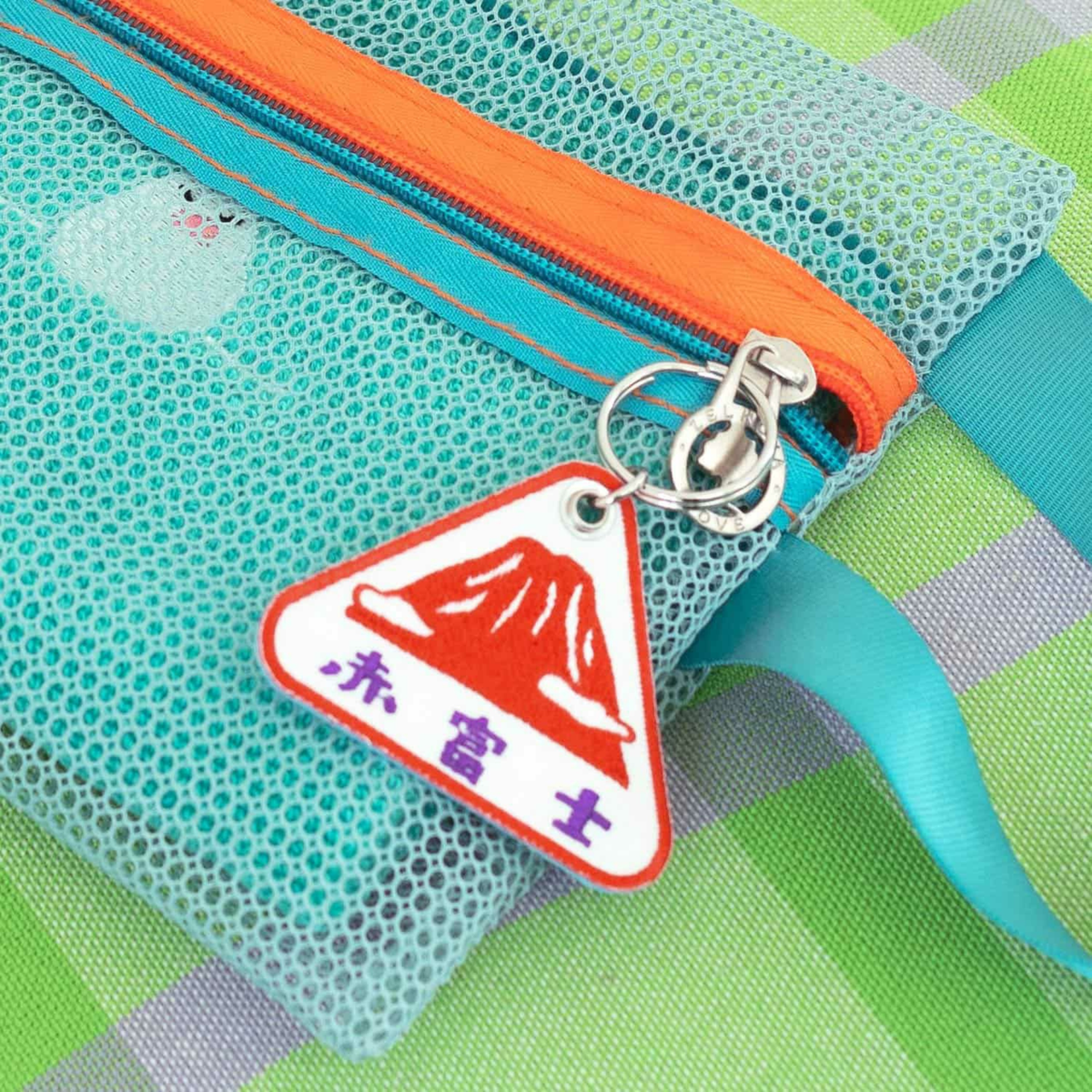 Een close-up van een turquoise tas van gaas met oranje rits en blauw lint, met de Hightide Stitch Work Sleutelhanger Fuji-een driehoekige geborduurde sleutelhanger met een rode berg en Japanse tekst op een groen-blauw geruite achtergrond.