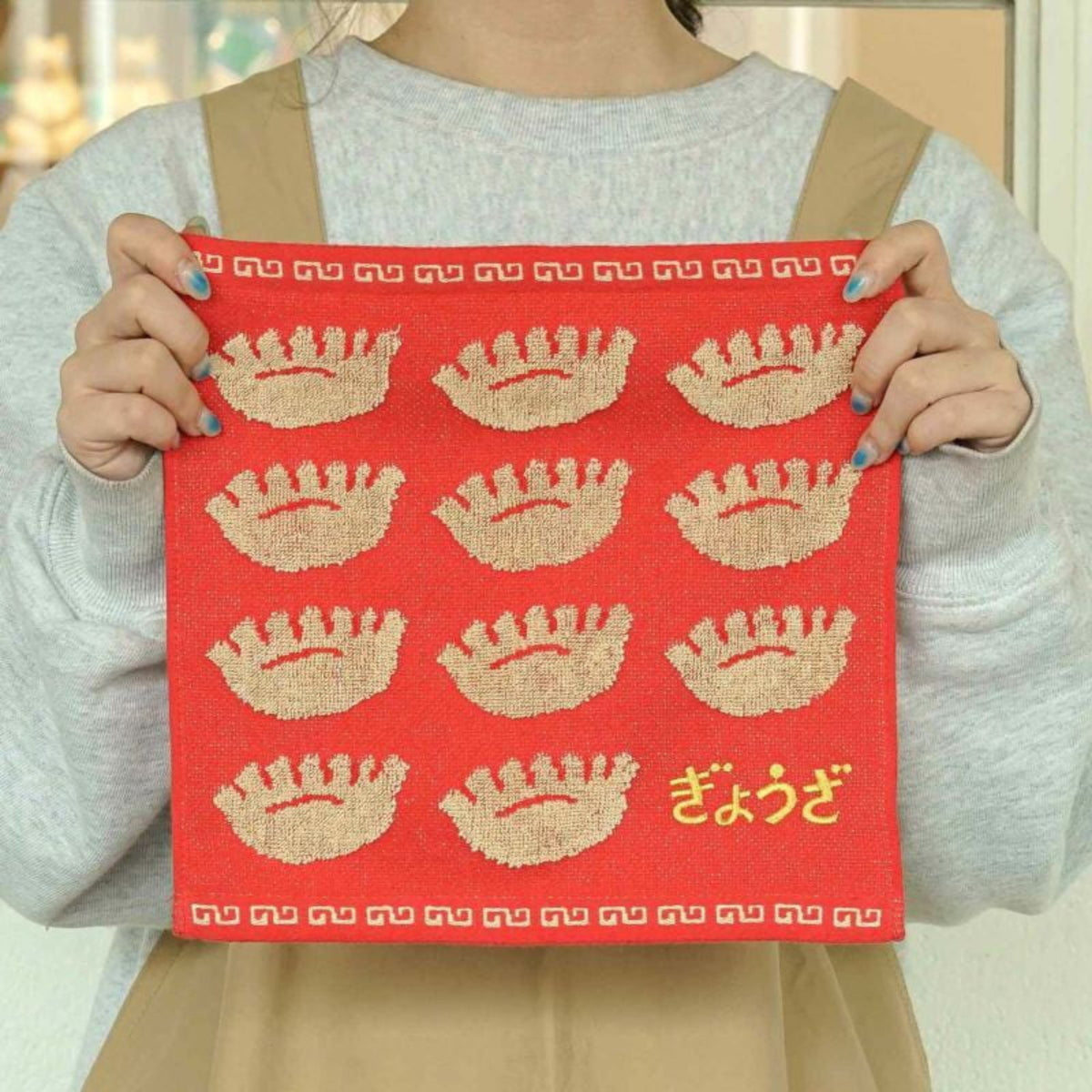Een persoon in een grijs shirt en beige schort houdt de Hightide Handdoek Dumpling 25/25 vast, een rode handdoek van Hightide met geborduurde dumplings, decoratieve randen en "gyoza" in het Japanse hiragana in de rechterbenedenhoek.