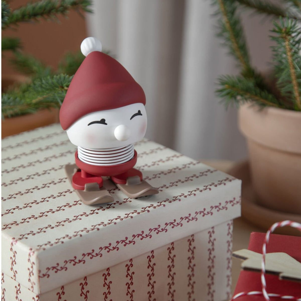 De Hoptimist Ski Bimble S Berry van Hoptimist - een klein, lachend beeldje met een rode muts en ski's - zit bovenop een geschenkdoos met een rood patroon. Het is een origineel geschenk en een perfecte winterdecoratie met dennentakken op de achtergrond.