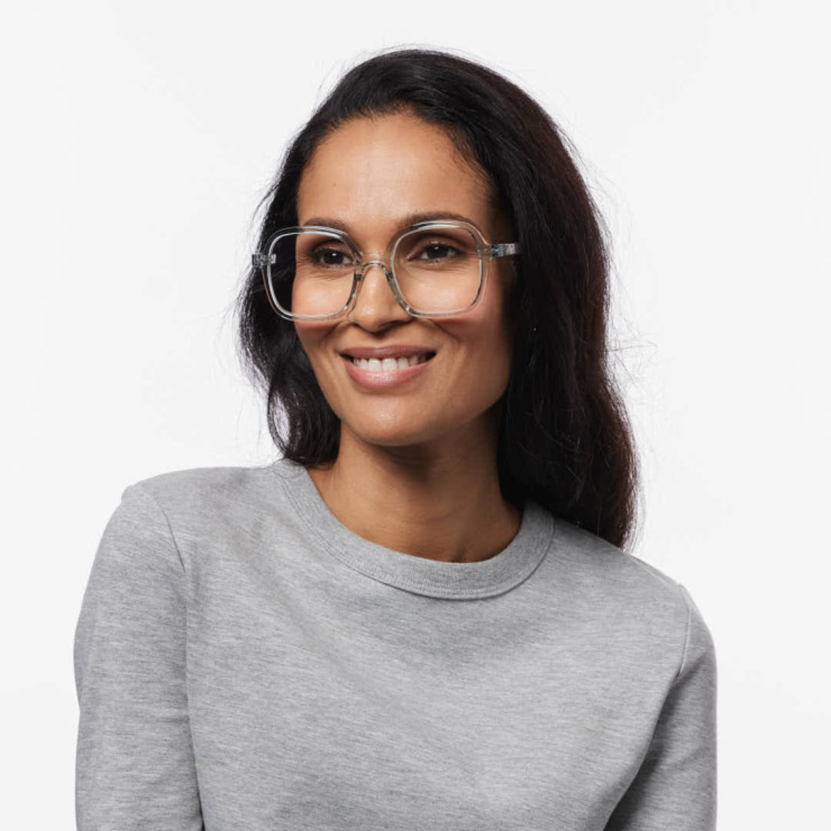 Een vrouw met lang donker haar draagt een oversized, doorzichtige Izipizi Leesbril Office Frozen Blue met een blauwlichtfilter; ze glimlacht in een lichtgrijs sweatshirt tegen een effen witte achtergrond.