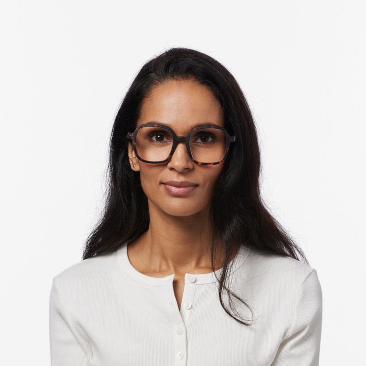 Een vrouw met lang donker haar draagt een grote, ronde Izipizi Leesbril Office Tortoise-bril en een wit overhemd met knopen; ze kijkt recht voor zich uit met een neutrale uitdrukking op haar gezicht, tegen een effen witte achtergrond.