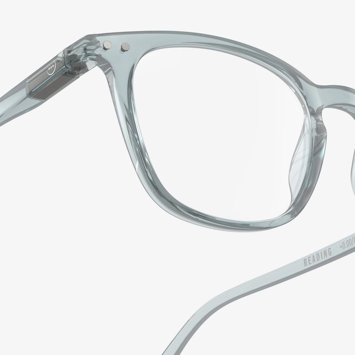 De Izipizi Leesbril #E Frozen Blue heeft een helder, transparant montuur met een glanzende afwerking. Op de arm staat "READING +0.00", waardoor het ideaal is voor mensen die last hebben van presbyopie. Van dichtbij afgebeeld tegen een witte achtergrond.