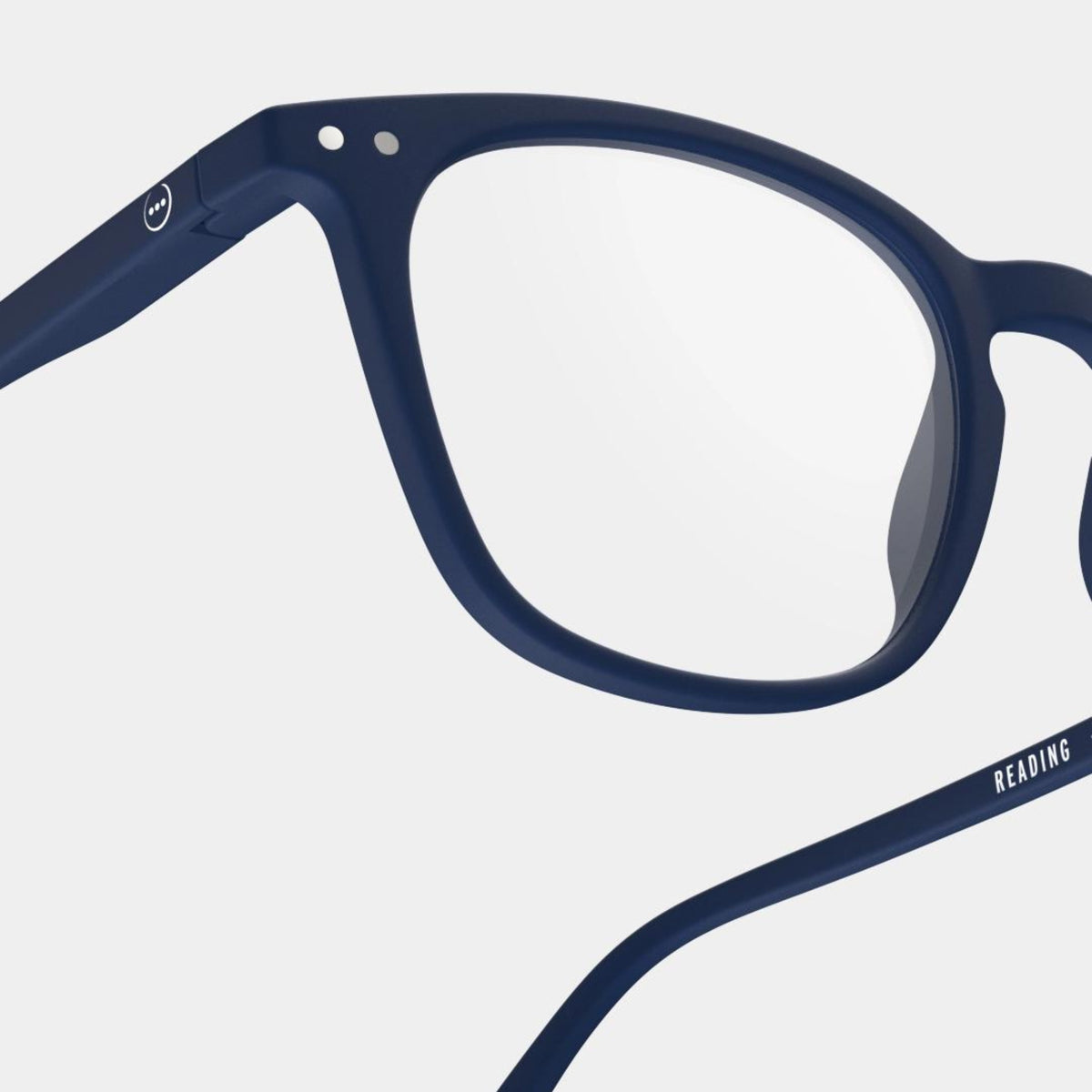 Een close-up van de Izipizi Leesbril #E Navy toont het ronde montuur en één volle lens, met marineblauwe veren met kleine metalen accenten en "READING" gedrukt aan de binnenkant van één arm, tegen een lichtgrijze achtergrond.