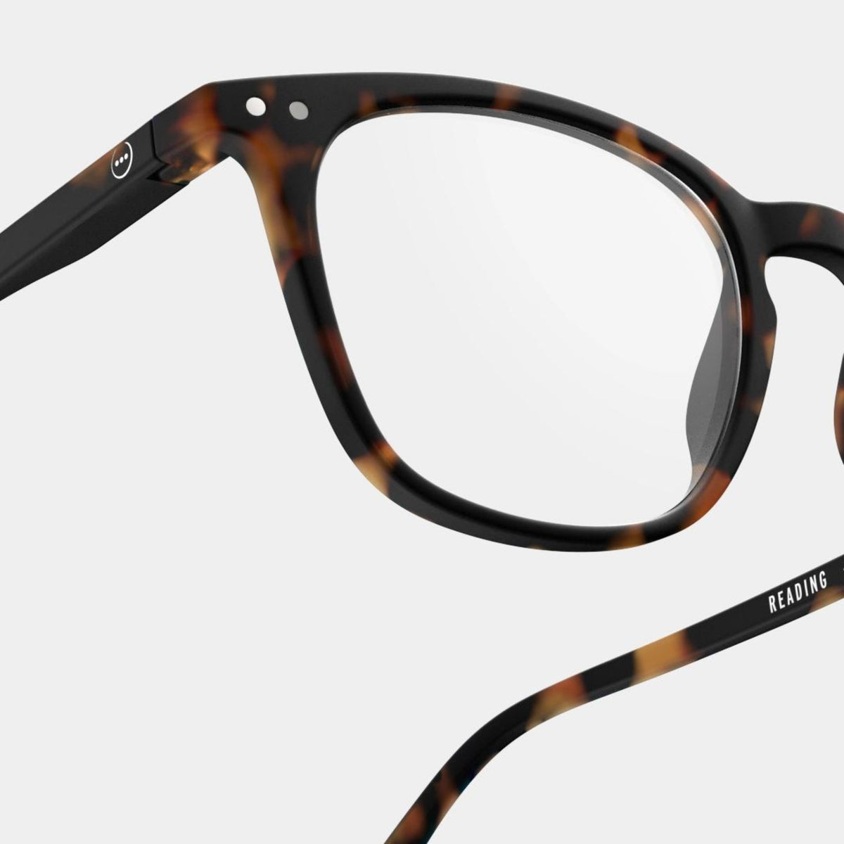 Close-up van de Izipizi Leesbril #E Tortoise leesbril van Izipizi met heldere glazen op een effen achtergrond. "READING" staat aan de binnenkant van het pootje en een witte smiley siert het scharnier. Stijlvolle unisex leesbril voor dagelijks gebruik.