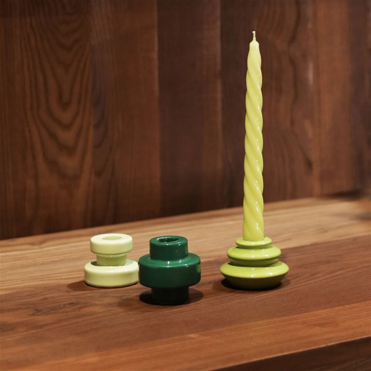 Drie ronde &Klevering Bravo Kaarsen Groen kandelaars van &Klevering, in lichtgeel en groen, zitten op hout; één houdt een gedraaide lichtgele kaars vast, de andere zijn leeg. Ideaal voor kleurrijke kaarsen of om cadeau te geven in een luxe geschenkdoos.