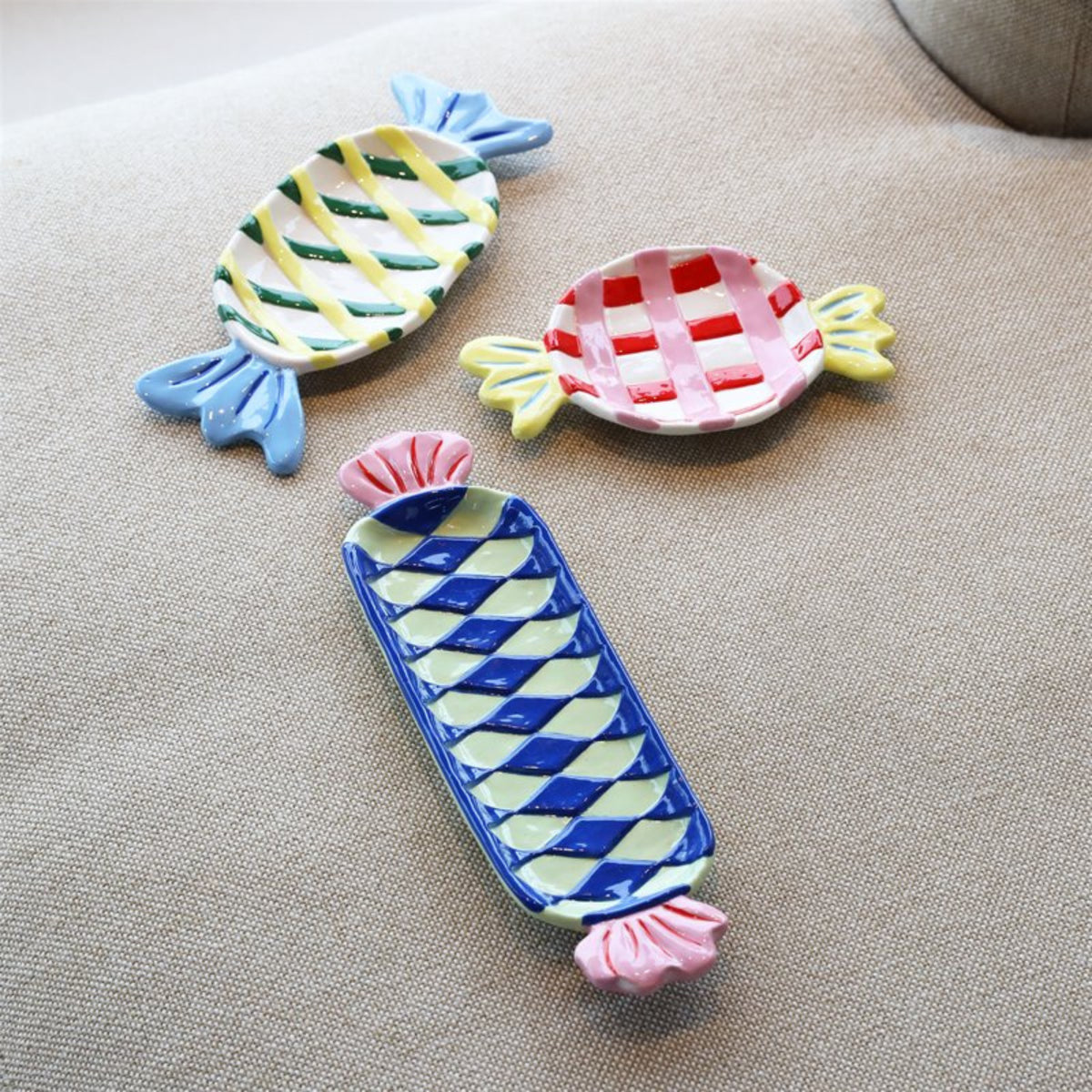 Drie &Klevering Goody Bord 27/11 keramische schalen van &Klevering, in de vorm van ingepakte snoepjes met rode, blauwe, gele en groene ruitjespatronen, staan op beige stof - perfect als speels middelpunt.