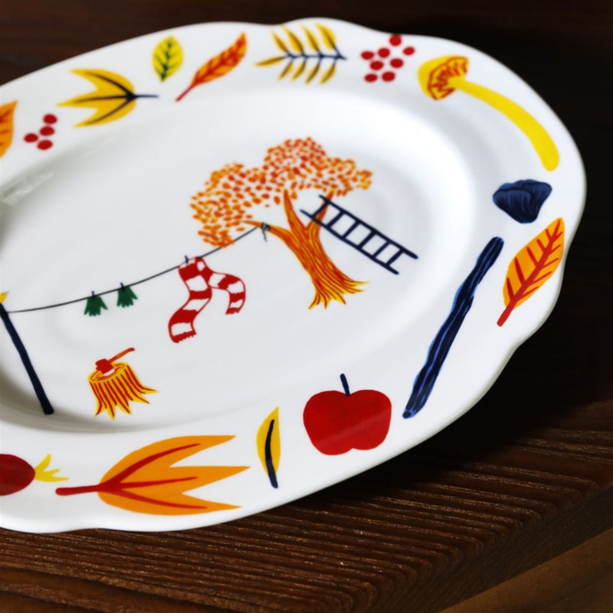 Het &Klevering Haines Schotel 28/22, een wit porseleinen bord van &Klevering met herfstblad- en appelillustraties en speelse details, rust op een donker houten oppervlak - ideaal voor cadeauverpakking of display.