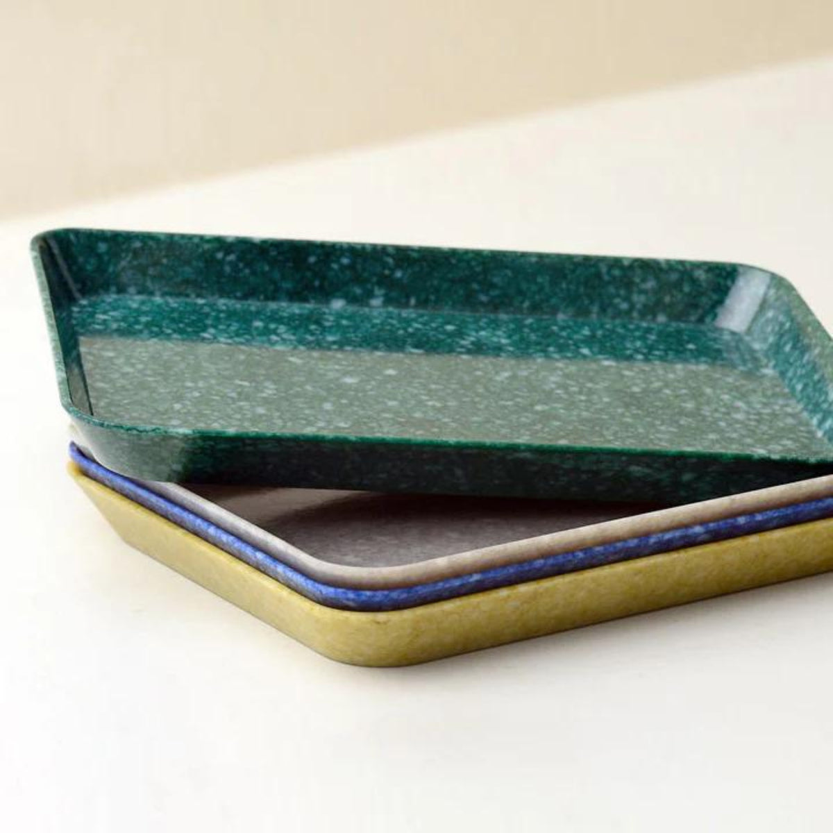 Drie rechthoekige dienbladen zijn gestapeld op een wit oppervlak. Bovenop staat het Melamine Marbled Dienblad Navy 23/16 van Melamine Marbled, met een groen gespikkeld patroon. Het middelste dienblad is grijs met een blauwe rand en het onderste is olijfgroen - ideaal voor kantoorgebruik.