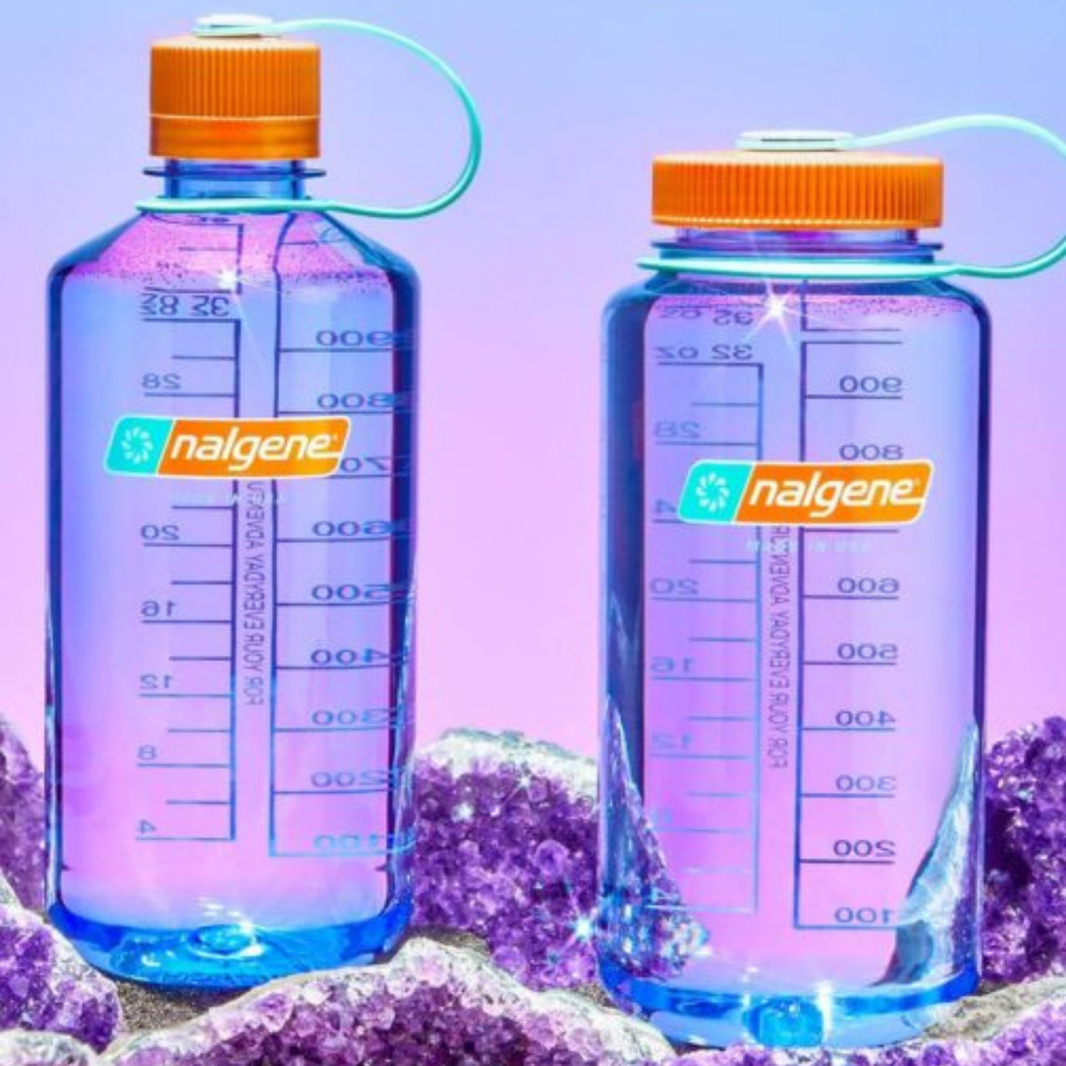 Twee Nalgene Drinkfles Brede Opening Amethyst 1L staan op paarse kristallen, tegen een roze-paarse achtergrond. De BPA-vrije flessen tonen maatstrepen, het Nalgene-logo en hebben oranje deksels.