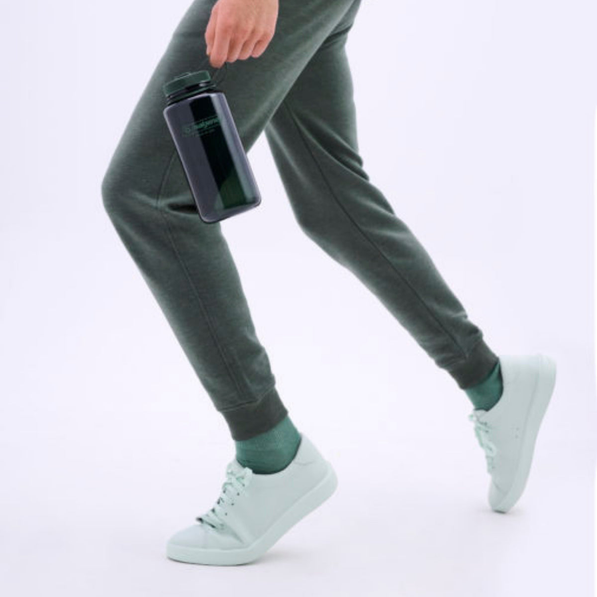 Persoon in groengrijze joggingbroek en mintgroene gympen loopt met een Nalgene Drinkfles Brede Opening Jade 1L waterfles aan zijn zijde. Alleen het onderlichaam en de hand zijn te zien tegen een lichte achtergrond. Merk: Nalgene.