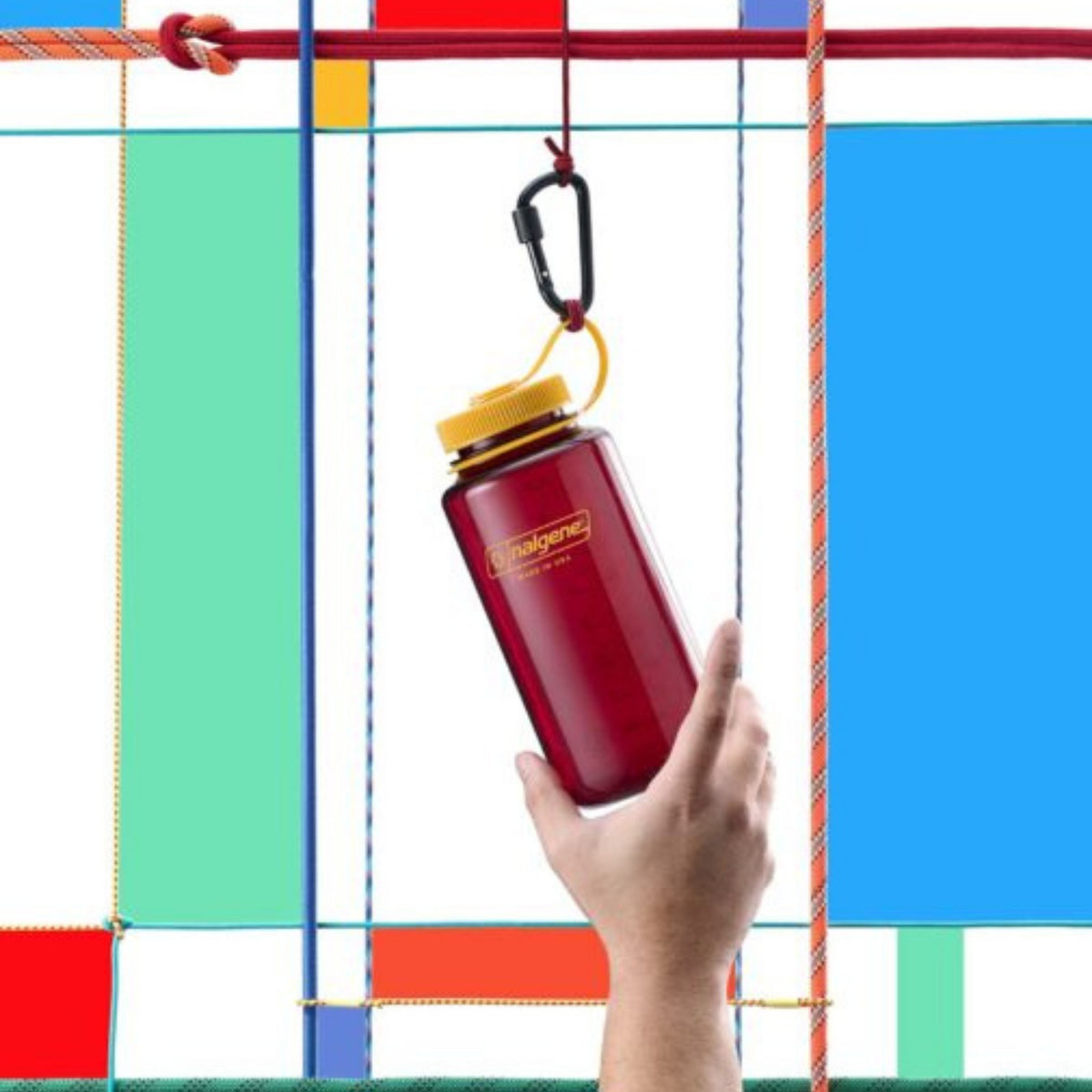 Een hand houdt een rode Nalgene Drinkfles Brede Opening Laker 1L met gele dop, bevestigd aan een karabijnhaak. Op de achtergrond zijn kleurrijke geometrische vormen in blauw, rood, groen en wit zichtbaar.