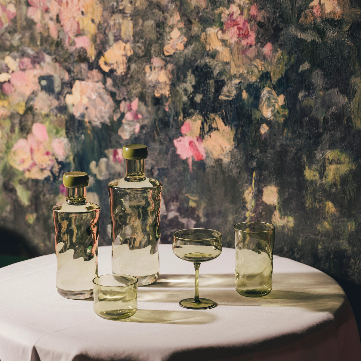 Drie Paveau Swirl Waterflessen Moss Groen 0,75 l en drie groene glazen staan op een rond wit tafelkleed, met op de achtergrond een vaag bloemenschilderij in roze, gele en groene tinten voor een duurzaam tintje.