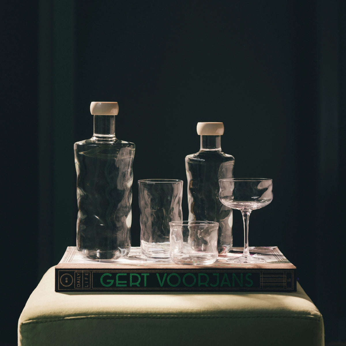 Twee Paveau Swirl Waterfles Wave Helder 0,75 l flessen met witte doppen, drie lege glazen en een coupeglas staan bovenop een groot boek met de titel Gert Voorjans op een lichtgroen oppervlak tegen een donkere achtergrond.