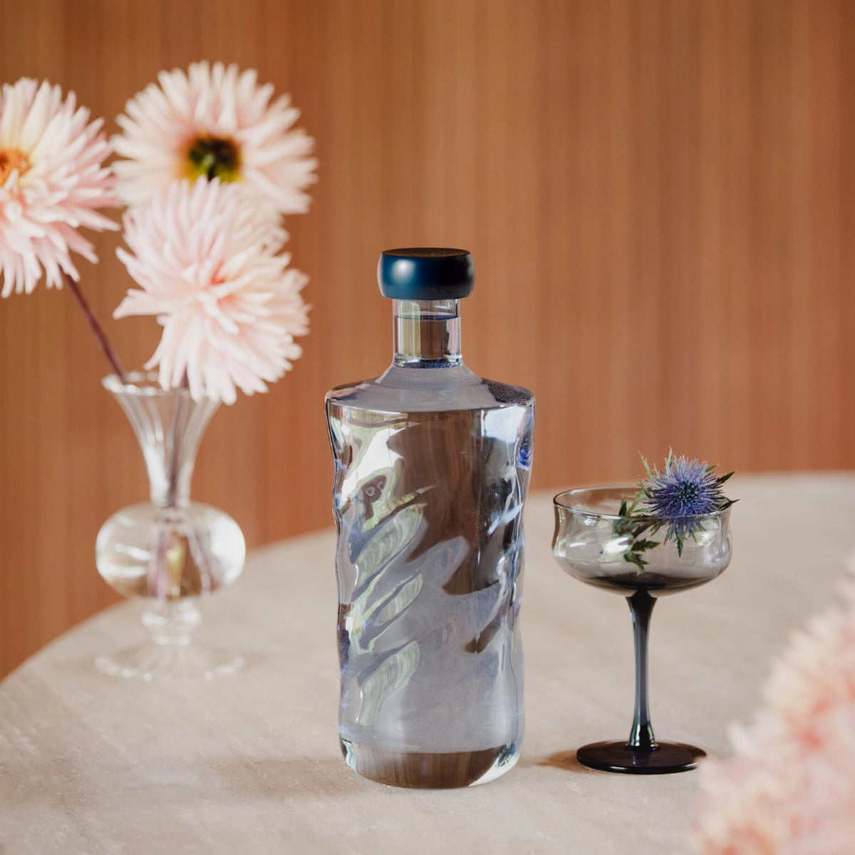 De Paveau Swirl Waterfles Midnight Blauw 1,25 l van Paveau staat op een ronde tafel naast een zwartgesteeld cocktailglas met blauwe distel en een vaas bleekroze bloemen, met een houten achtergrond.