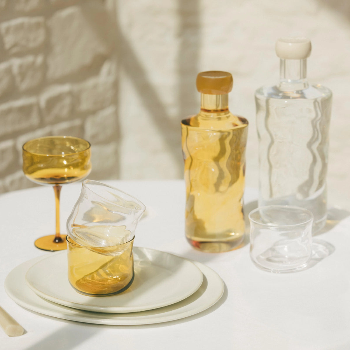 Op een zonovergoten tafel staat de Paveau Swirl Champagne Coupe Dune Geel 26 cl van Paveau, gecombineerd met amberkleurig en helder borosilicaatglaswerk, flessen, bekers en witte borden op een wit tafelkleed met zachte schaduwen.