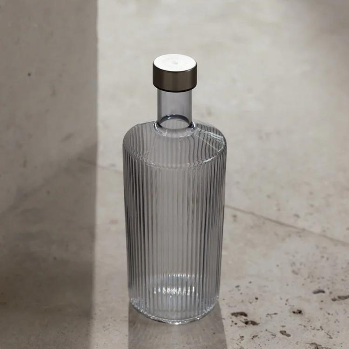 De Paveau Waterfles Twilight Blue 1,25 l van Paveau, met een helder geribbeld ontwerp en metalen kap, staat rechtop op een beige stenen oppervlak dat baadt in zacht natuurlijk licht.