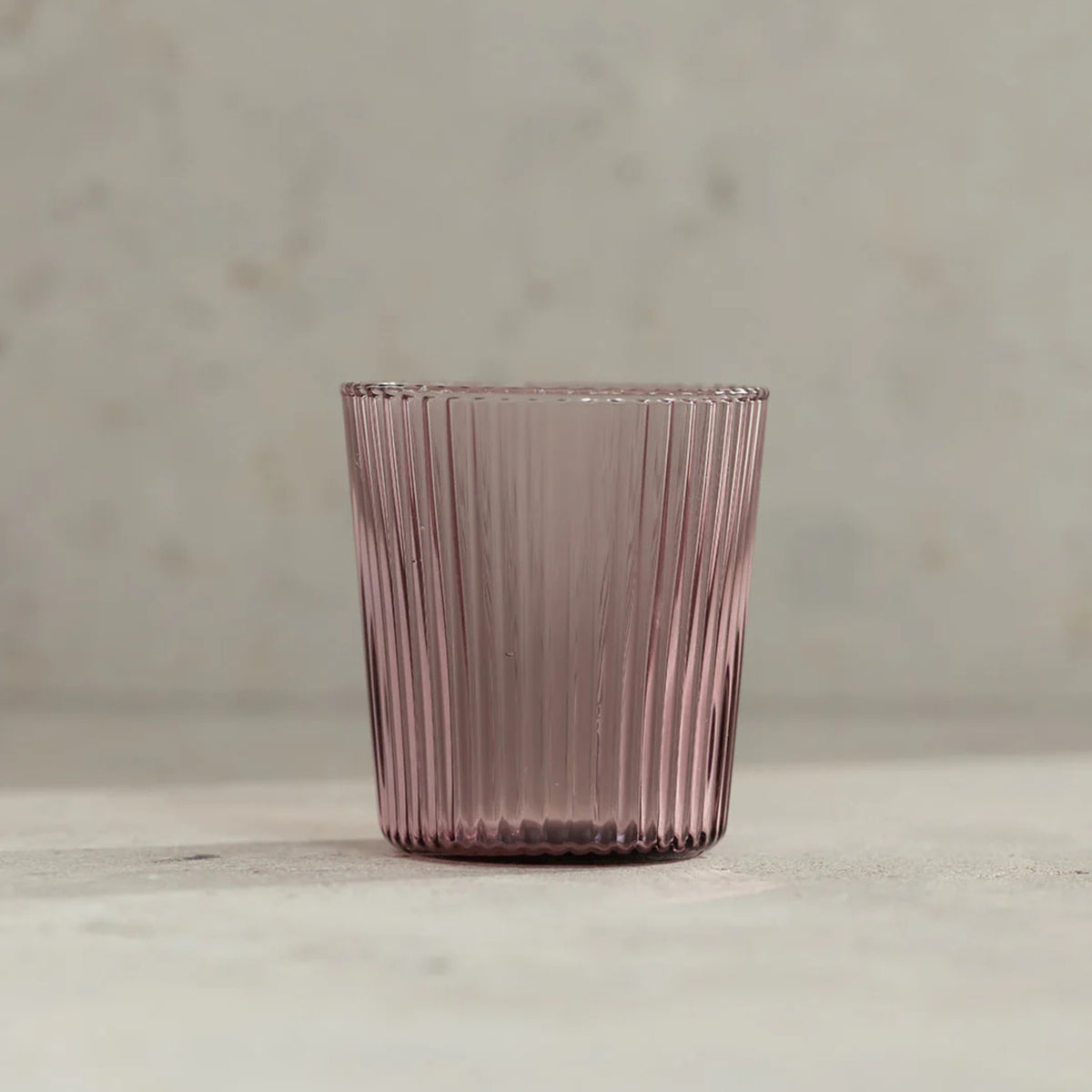 De Paveau Waterglas Broome van Paveau, met een roze tint en verticaal geribbeld ontwerp, staat op een zacht neutraal oppervlak - ideaal voor iedereen die op zoek is naar een stijlvolle, duurzame drinkbril.