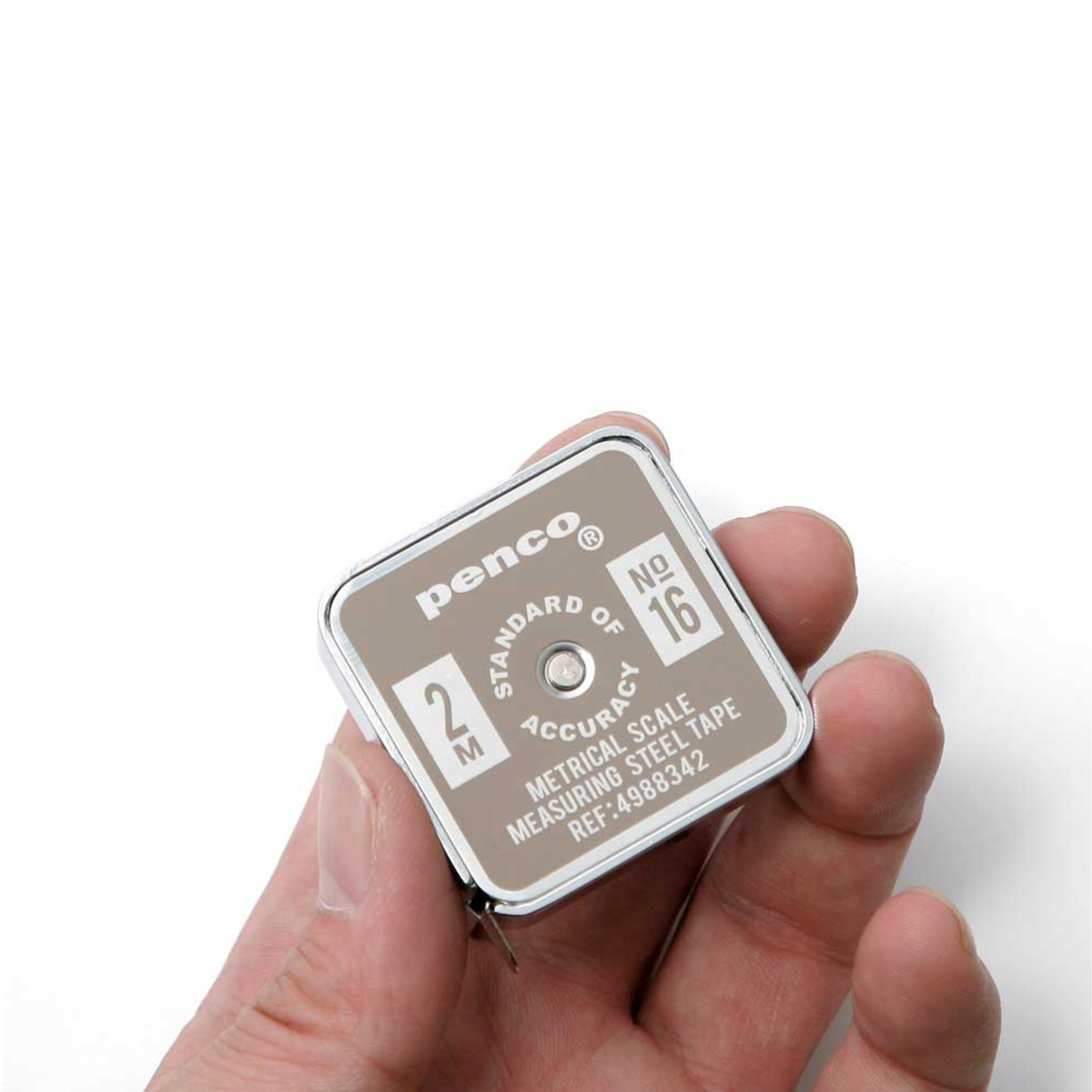 Een hand houdt een Penco Compacte Rolmaat Ivoor vast, een klein vierkant meetinstrument in zilver en beige met de opschriften 2M, STANDARD en NO 16, en productinformatie op de voorkant.