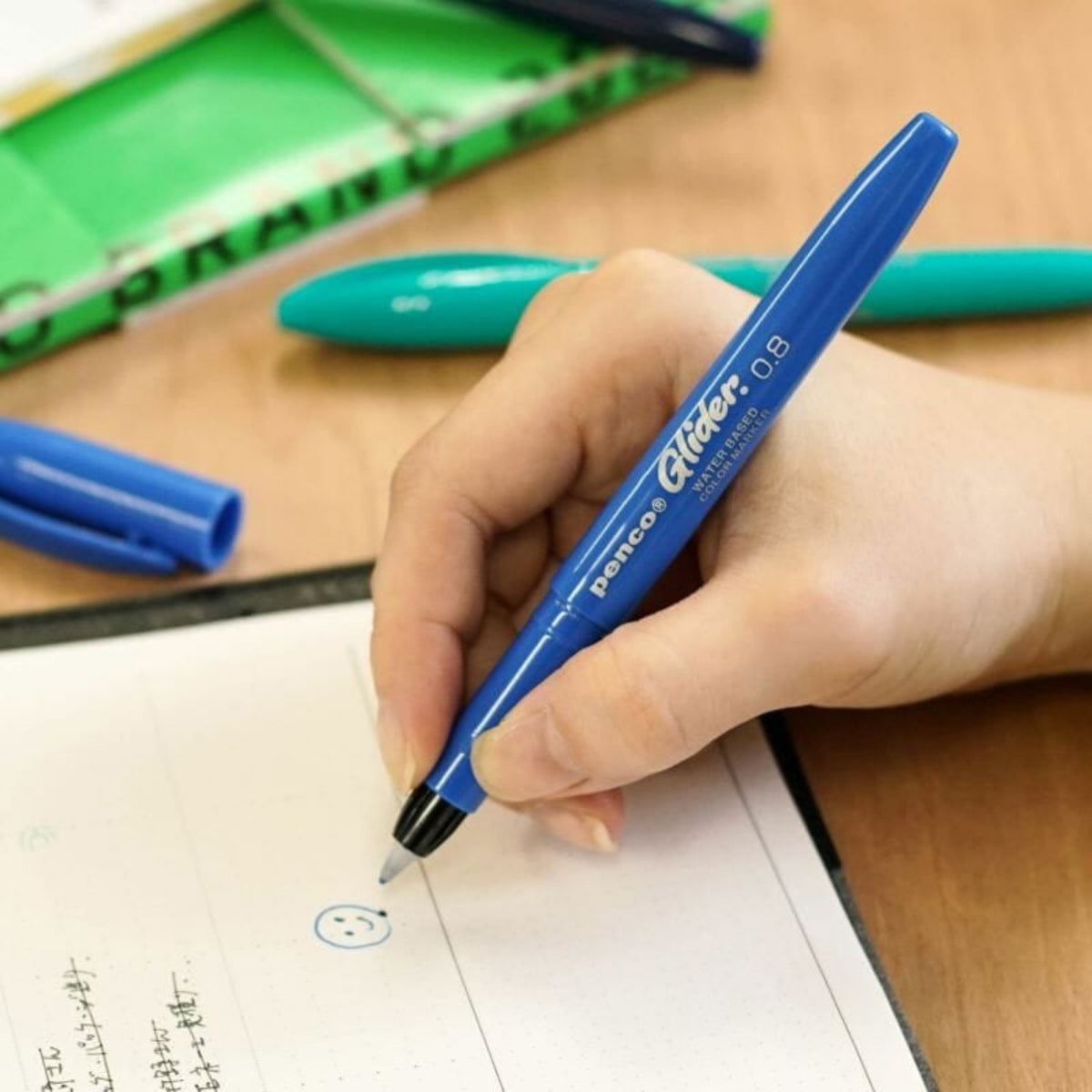Een hand houdt een Penco Glider Pen Blauw vast en tekent een smiley op gestippeld papier. Een open notitieboekje en andere blauwe en groene pennen van Penco liggen op het houten bureau en beloven een zijdezachte schrijfervaring.