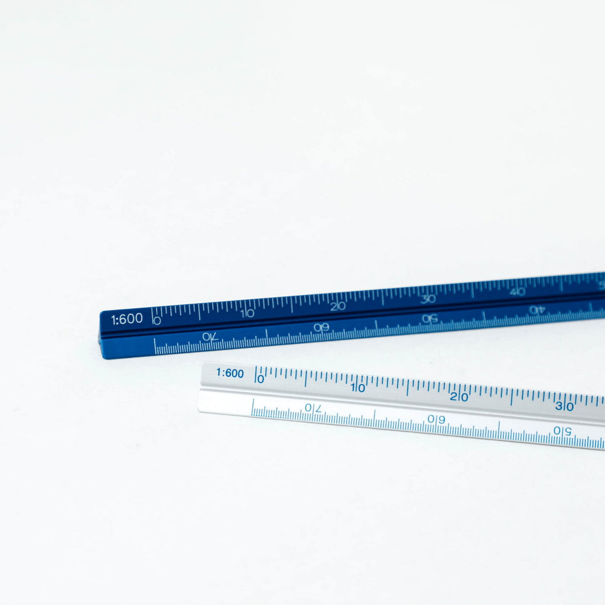 Twee driehoekige aluminium linialen van het merk Penco, type Lat Aluminium Zilver, liggen op een witte ondergrond; één blauw met schaal 1:600 voor technische tekenen en één wit, beide met maatverdeling.